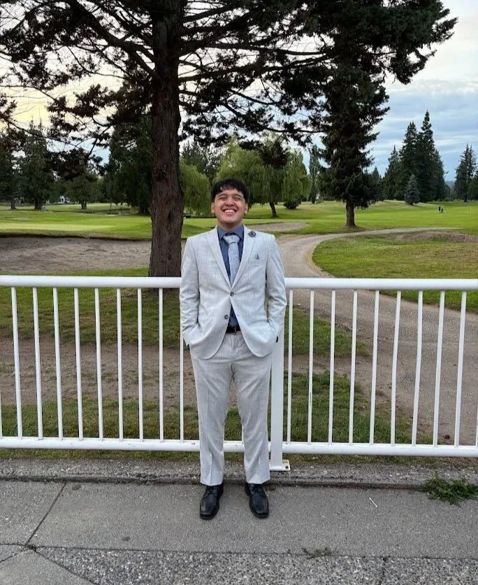 A young man in a light gray suit and black shoes, smiling and standing with hands in pockets in front of a white metal fence in a park with trees and grassy areas.