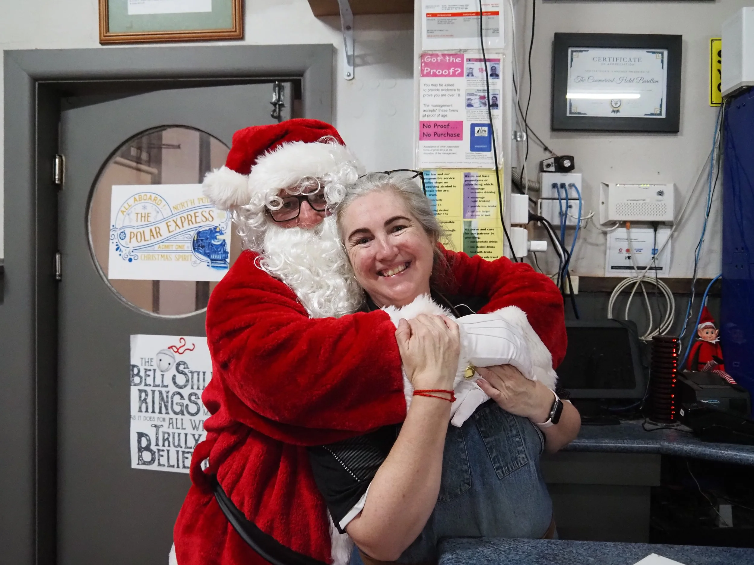 A person dressed as Santa Claus hugging a woman and smiling. They are indoors, with signs, a framed certificate, and electrical equipment visible on the wall behind them.