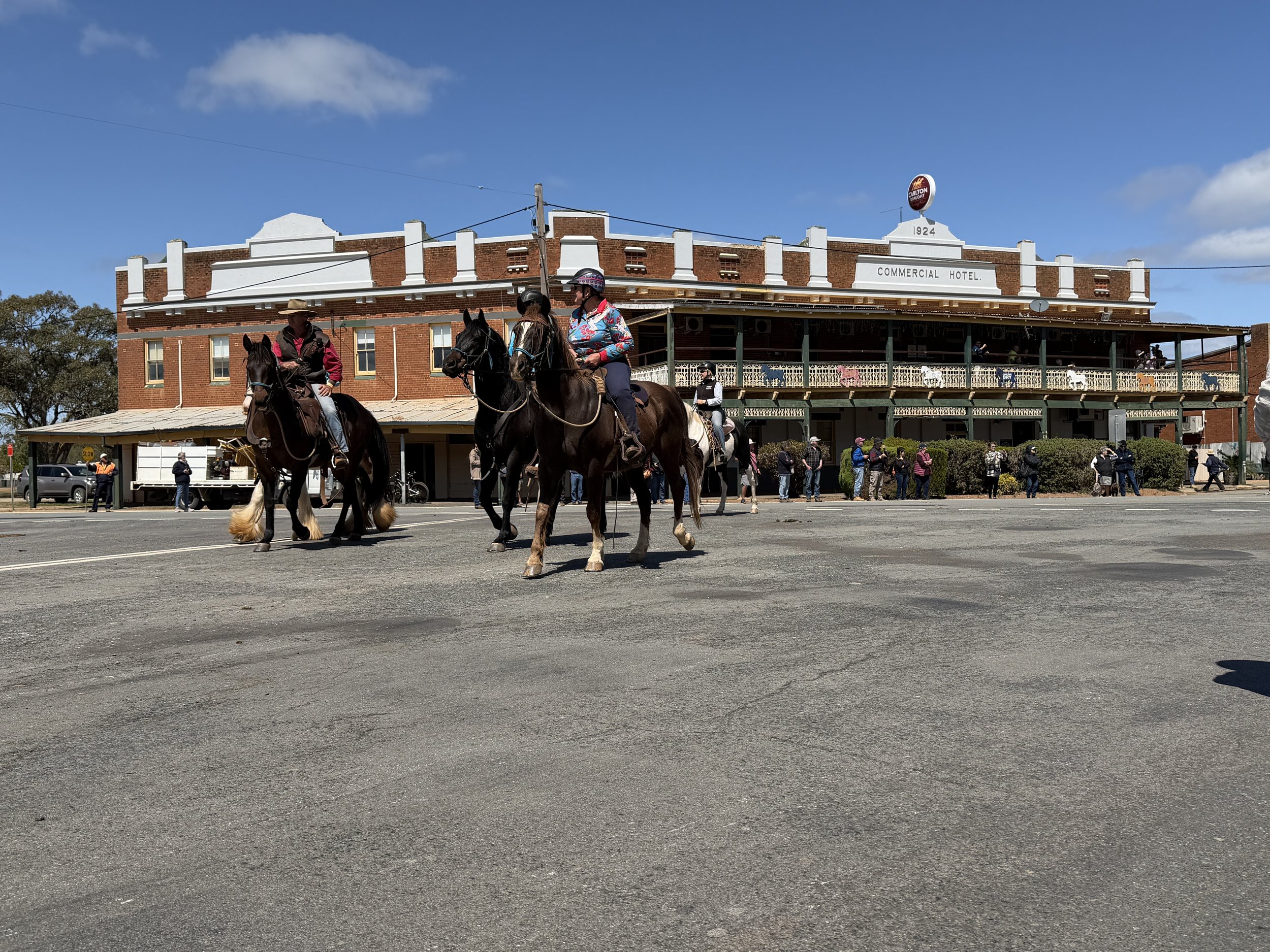 People riding horses in front of a historic brick hotel building on a sunny day.