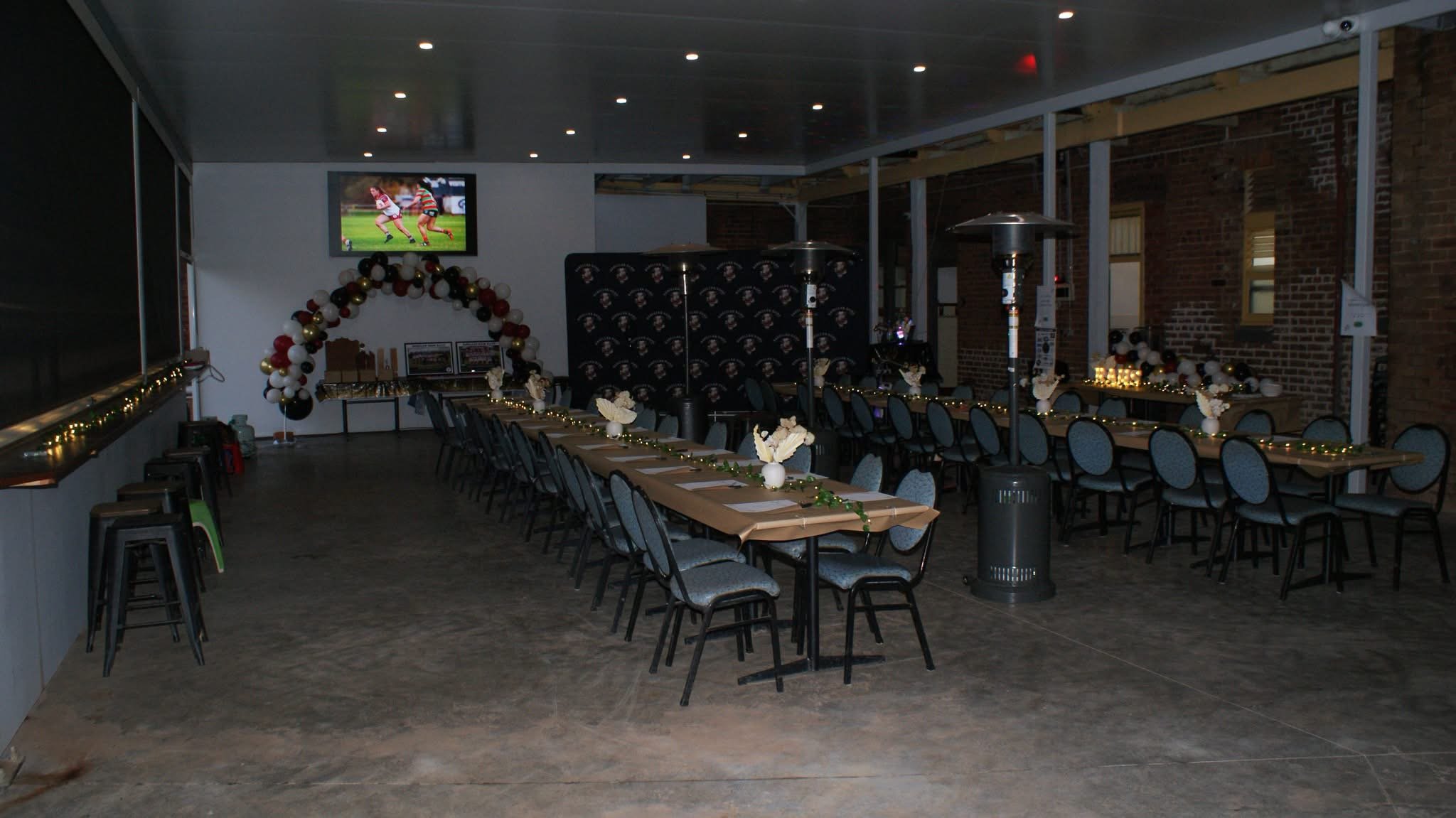 Indoor celebration space decorated with balloons, fairy lights, and tables set for an event, with a television displaying a sports game on the wall.