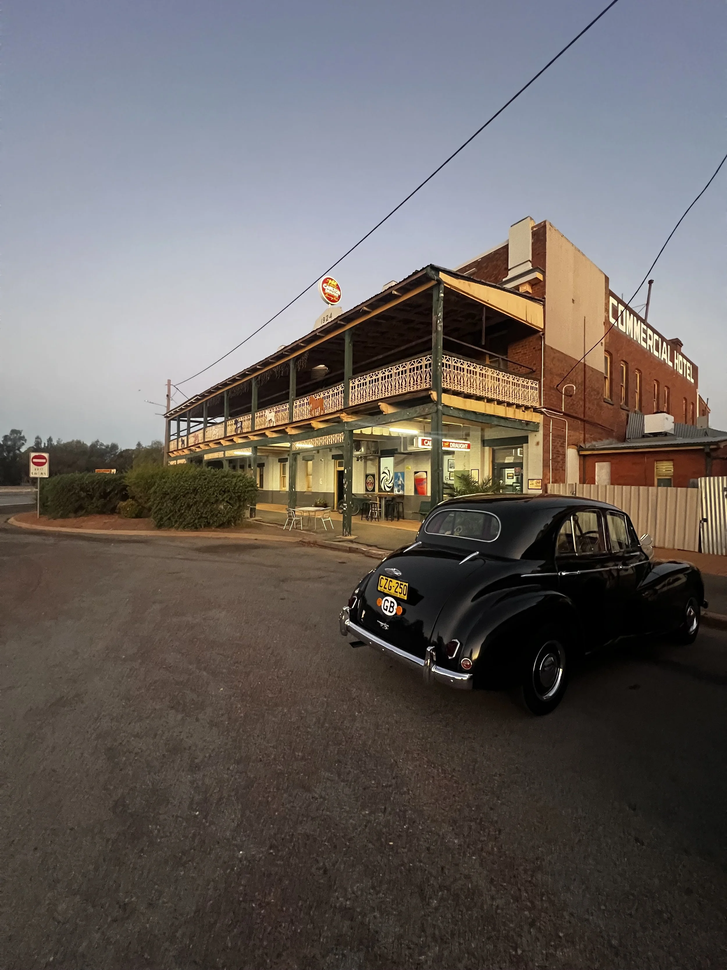 A black vintage car parked on the street in front of a building named 'Commercial Hotel,' with a balcony and brick exterior, under an evening sky.