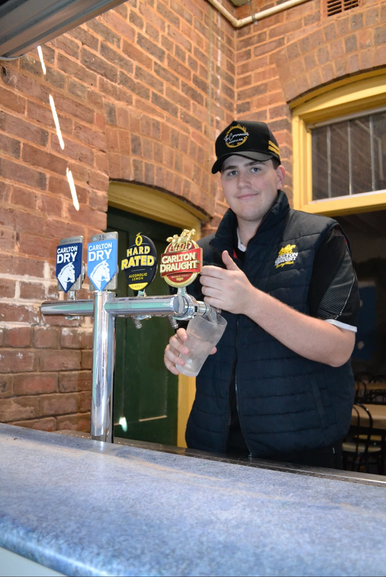 A young man wearing a black Carlton Draught jacket and cap standing at a bar with beer taps labeled Carlton Dry, rated, and Carlton Draught. He is holding a glass of beer and smiling.