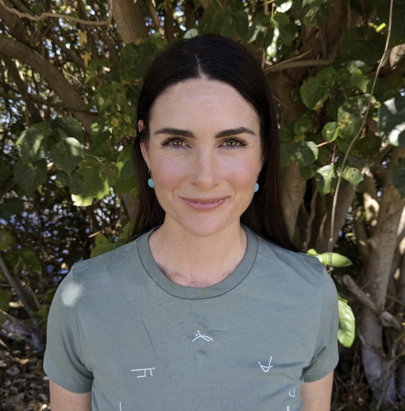 A woman with long dark hair, wearing turquoise earrings and a green T-shirt with a small logo, is smiling and standing outdoors in front of a leafy tree.