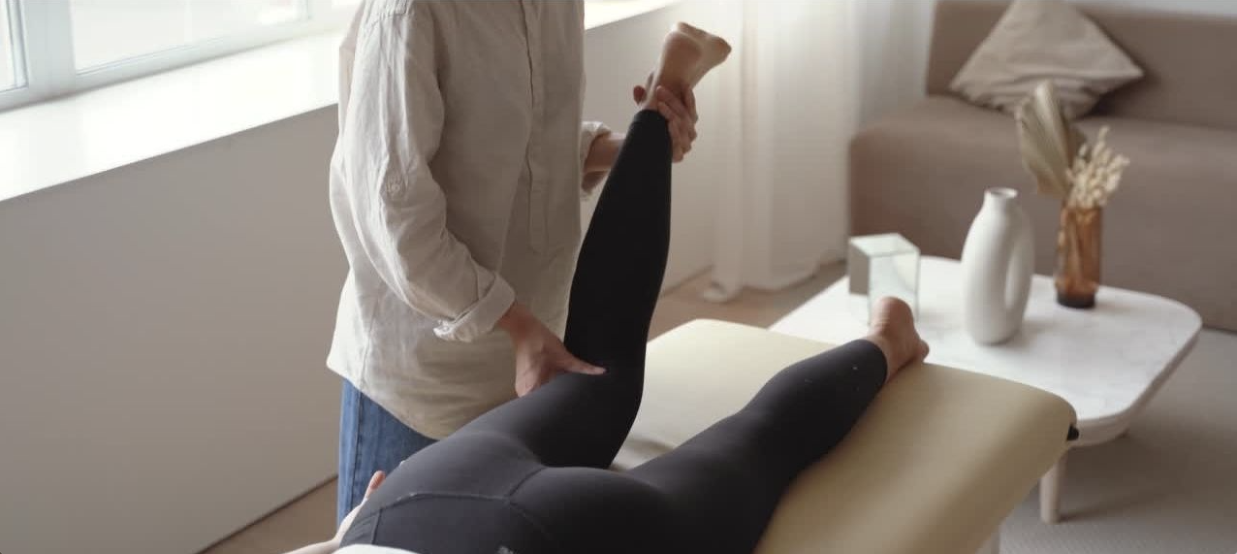 Person receiving physical therapy on their leg in a clinic, with a therapist holding their ankle and knee, and a side table with vases and decorative objects in the background.