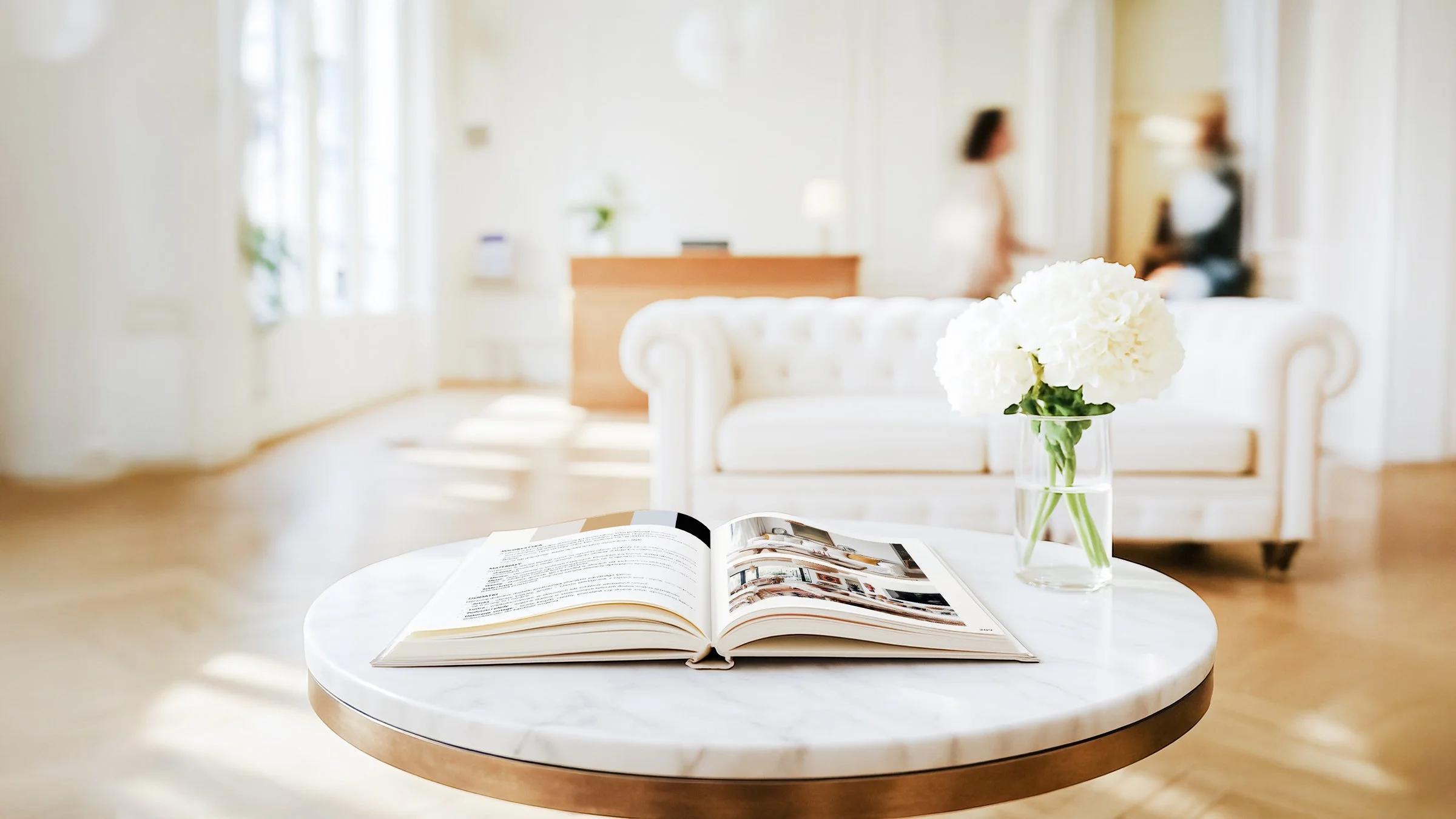 Open Homessence magazine on a white marble table with a vase of white hydrangeas, in a bright, modern living room with a white sofa and blurred figures in the background.
