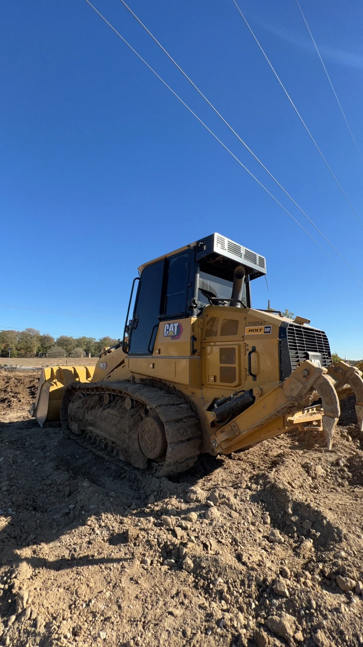 A yellow bulldozer with tracks on a dirt construction site under a clear blue sky with power lines overhead.