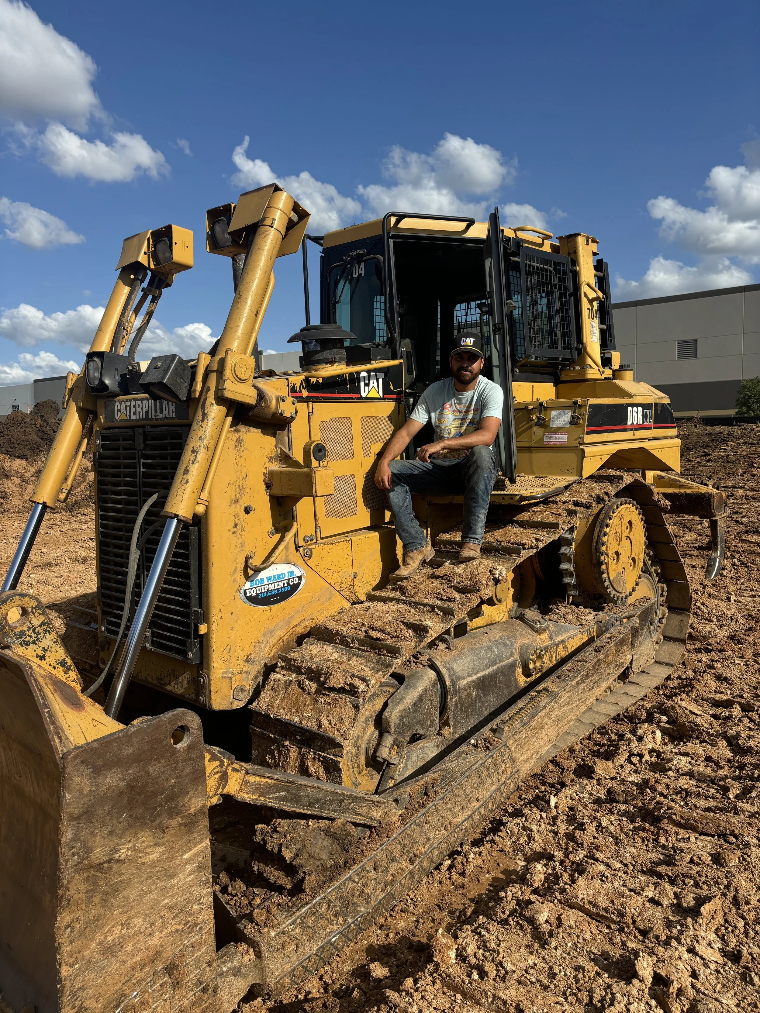 A man sitting on a yellow Caterpillar D6RXL bulldozer with a partially cloudy blue sky in the background. The bulldozer is on dirt and appears to be at a construction site.