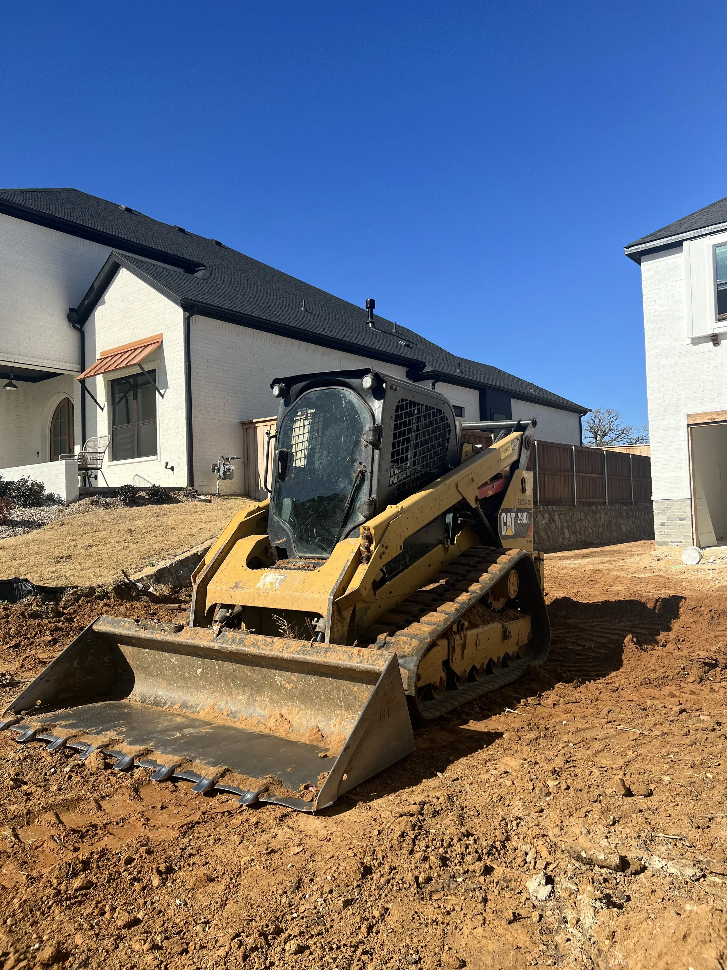 Construction site with a small yellow bulldozer on dirt ground, residential houses in the background under a clear blue sky.