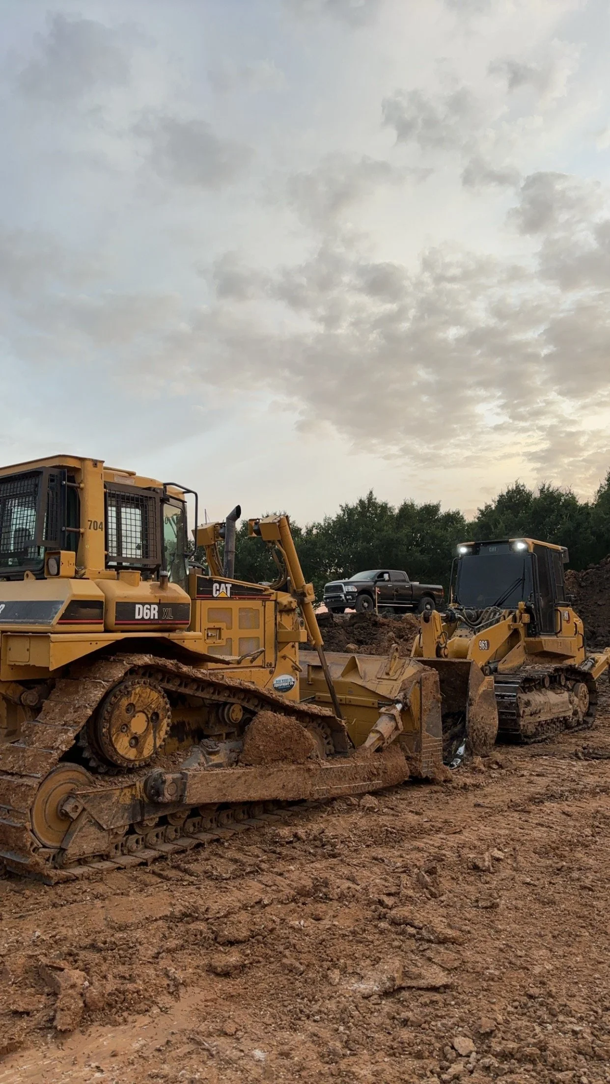 Construction site with bulldozer and excavator on dirt, truck on hill, cloudy sky.