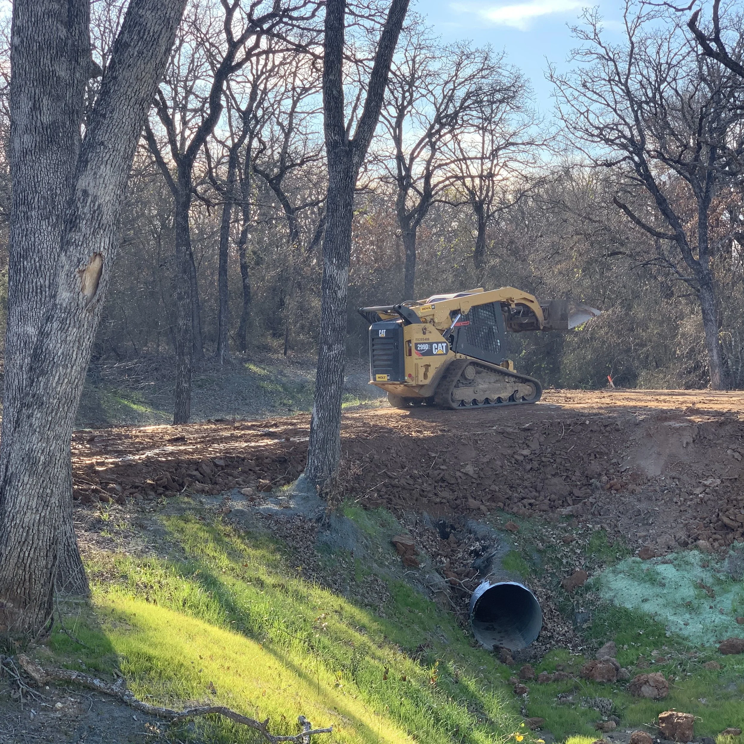 A construction site with trees, dirt, and a yellow Caterpillar excavator digging a hole, with a black pipe on the ground and a bright sky overhead.
