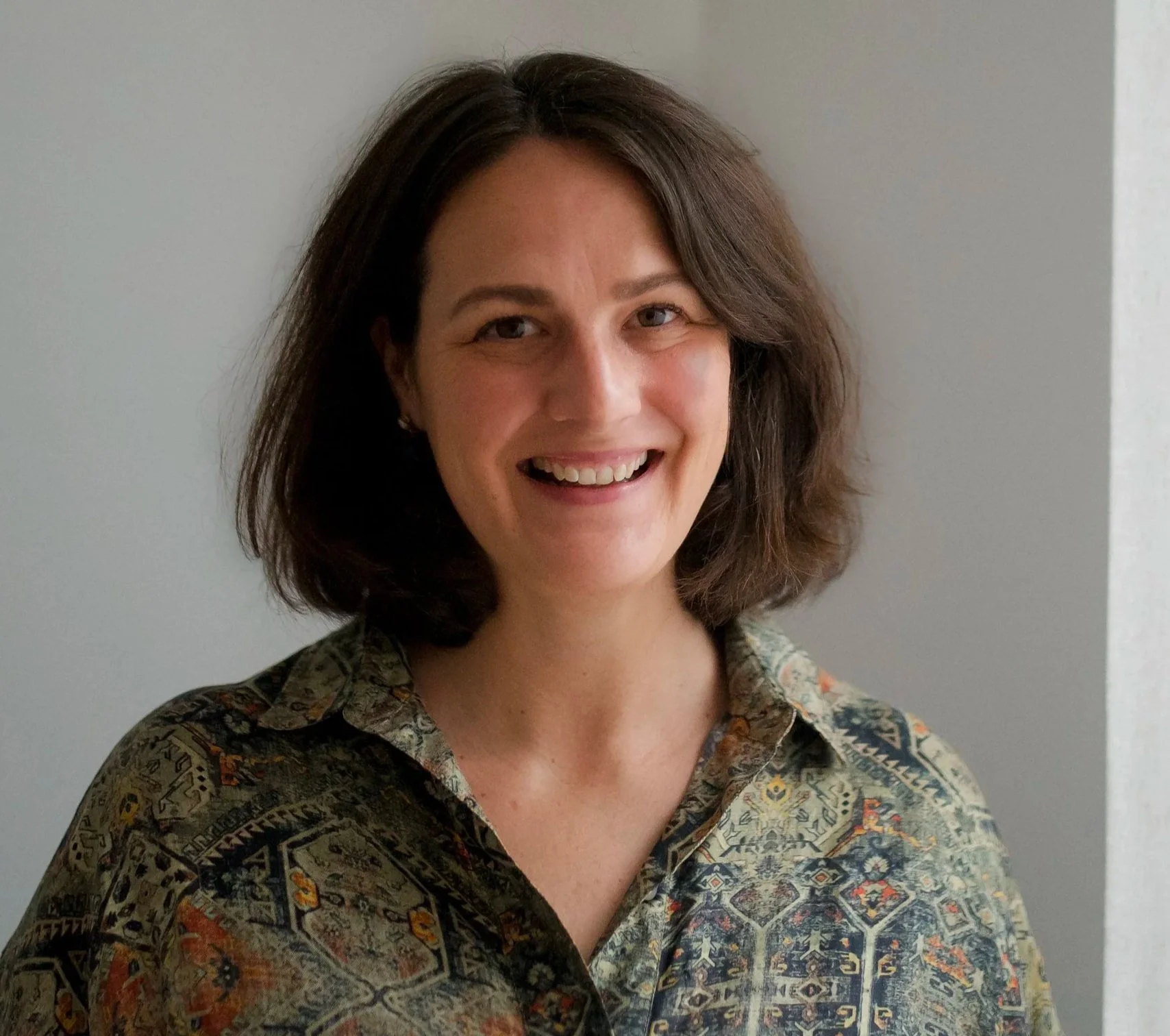 A woman with shoulder-length brown hair smiling, wearing a patterned button-up shirt, standing against a neutral wall.