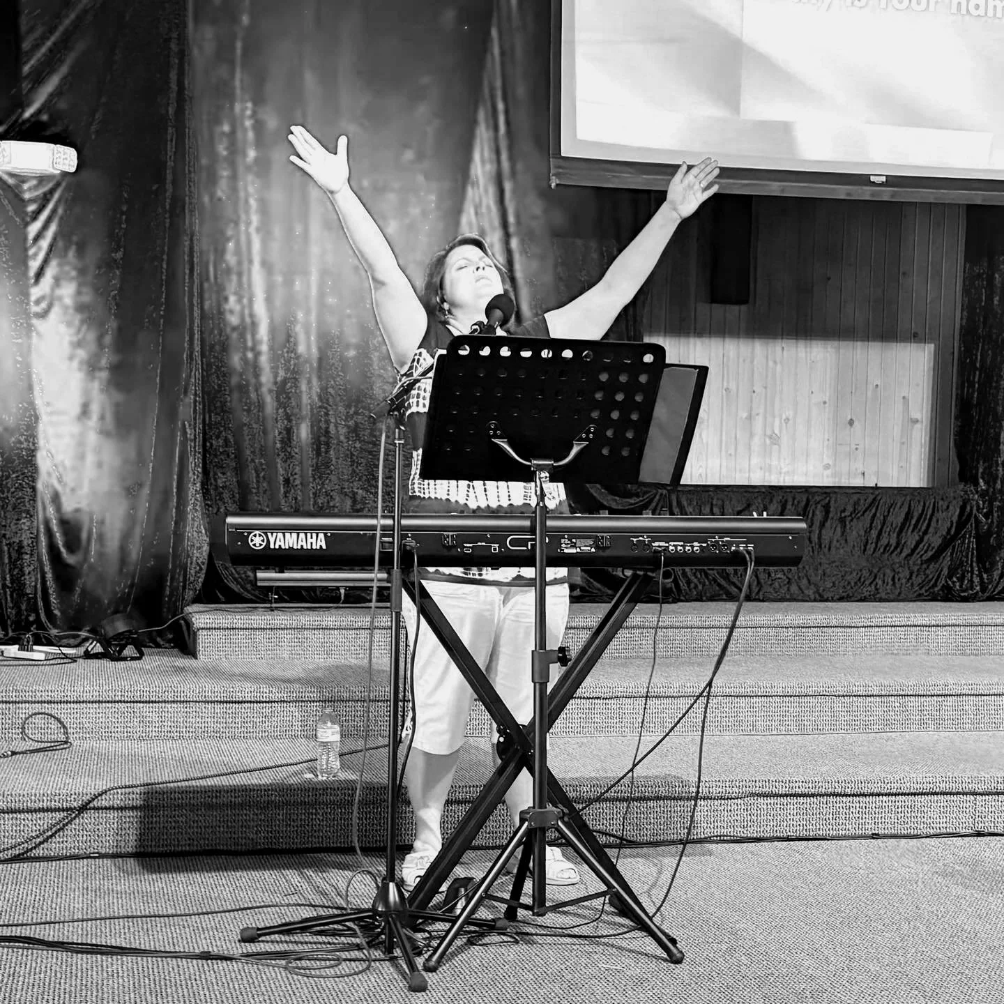 A woman stands on a stage with her arms raised and eyes closed, singing or praying, in front of a Yamaha keyboard, with music stand, microphone, and a projection screen behind her.