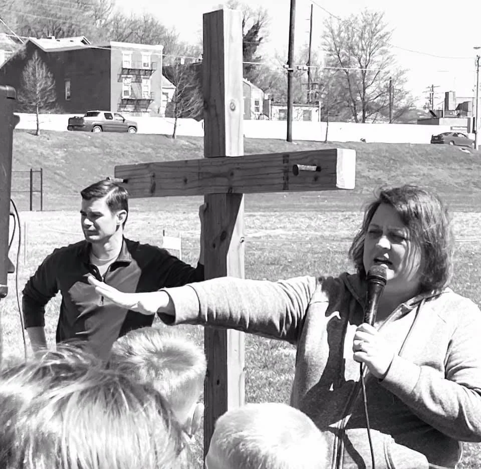 A woman with glasses holding a microphone and gesturing with her hand in an outdoor area, with a wooden cross structure behind her and a small crowd of people in the foreground. In the background, there are houses, a car, and trees.