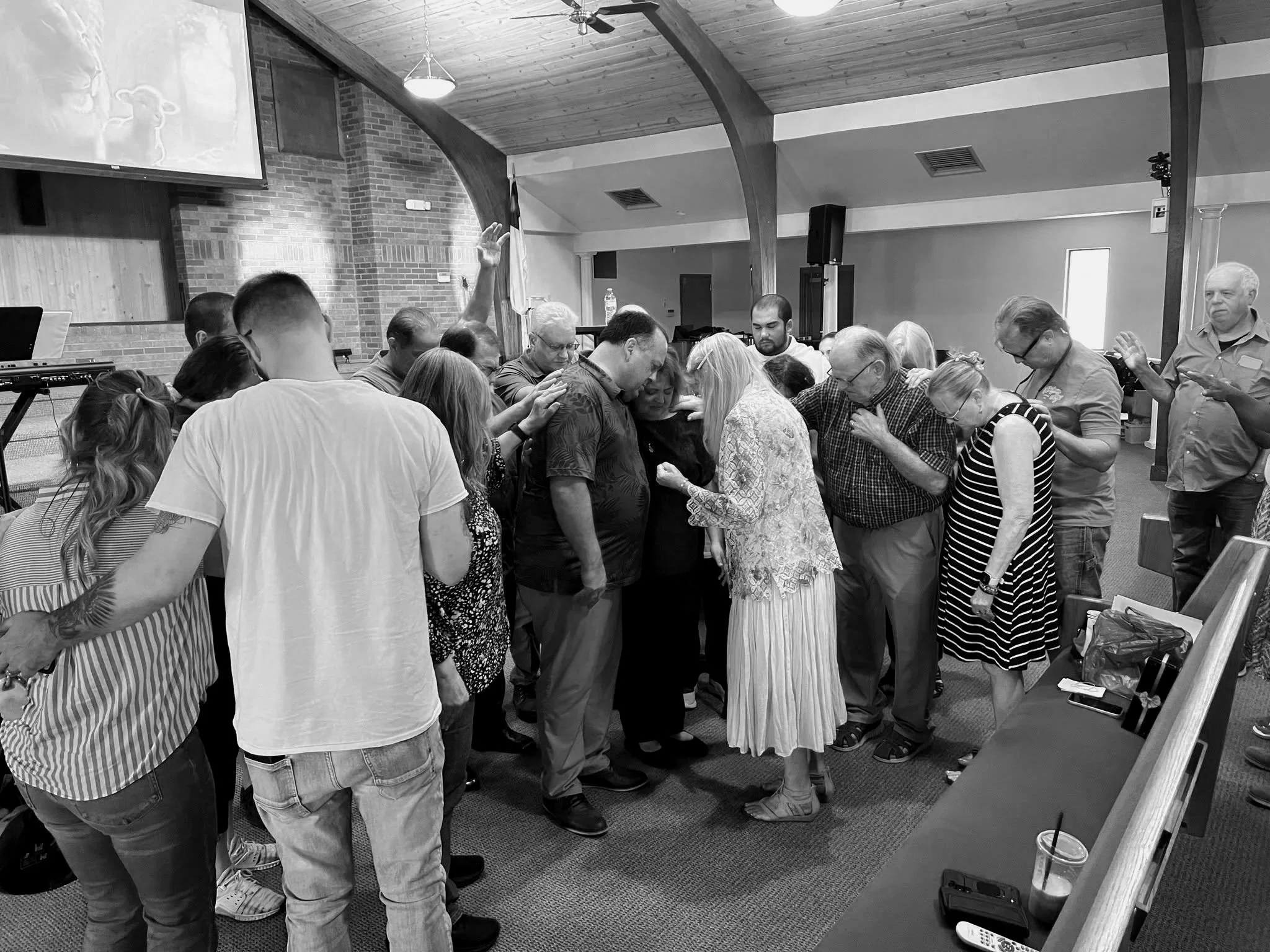 A group of people gather in a church or religious setting with their heads bowed, possibly in prayer, with one person raising their hand. The interior features wooden and brick walls, a large projection screen, and various electronic equipment.
