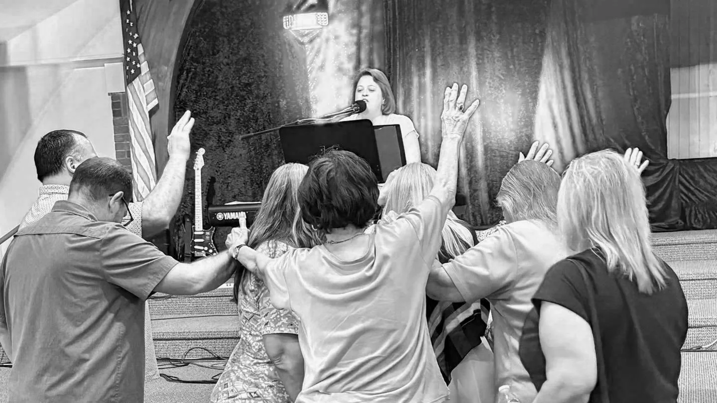 A woman singing at a microphone while playing a keyboard on stage. Several people in the audience are raising their hands, with some touching or holding hands. The background has a curtain, an American flag, and a guitar.