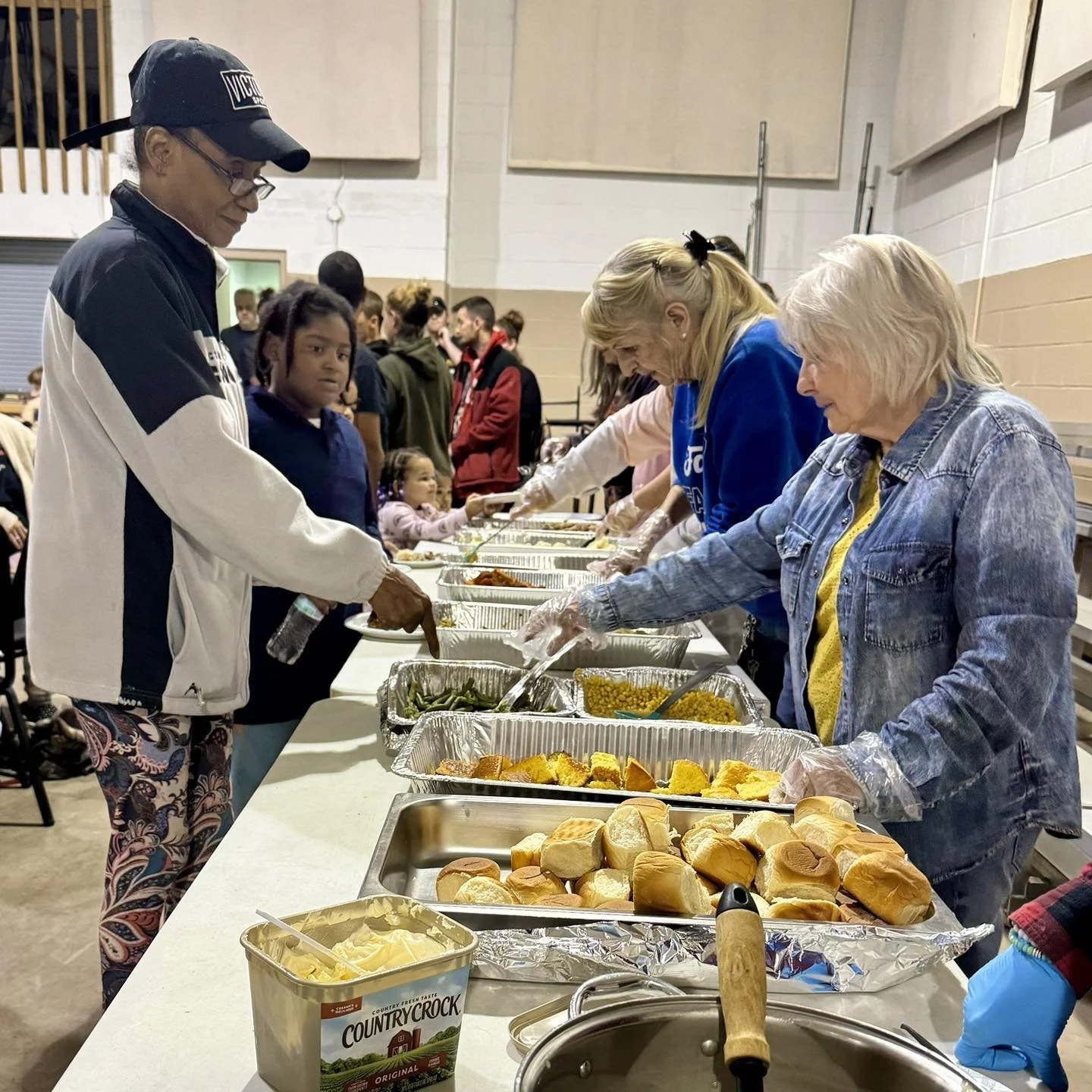 People serving food at a community meal event, with trays of bread, rolls, and various dishes on the table.