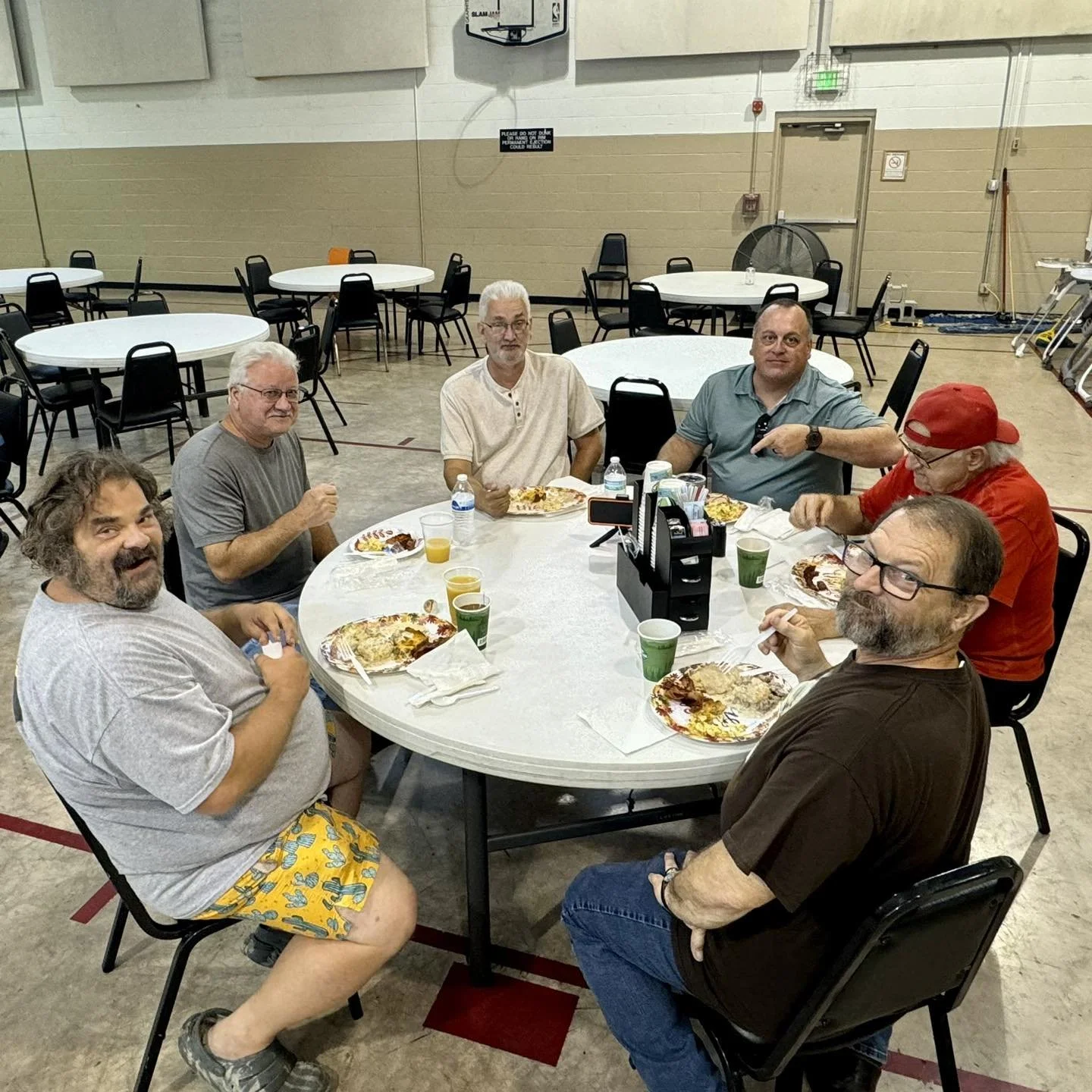 Six people sitting around a round table eating pizza and drinking juice in a large event room.