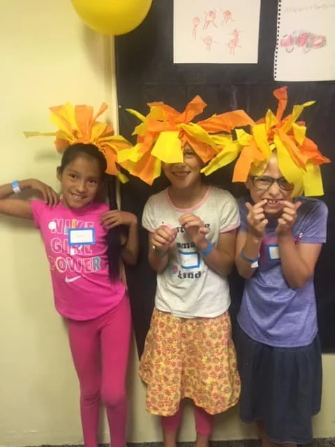 Three children wearing headbands made of large yellow and orange tissue paper flowers. They are standing in front of a wall with children's drawings and a blackboard, smiling and posing for the camera.