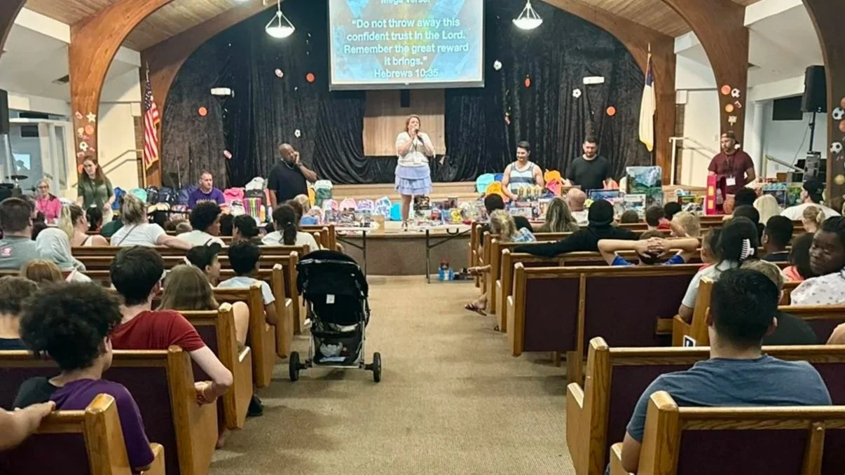 A congregation gathered in a church sanctuary, listening to a woman speaking on stage. The stage has various items displayed on tables, and a large screen above shows a Bible verse. The audience includes children and adults sitting on wooden pews, with some children in strollers and wearing headphones.