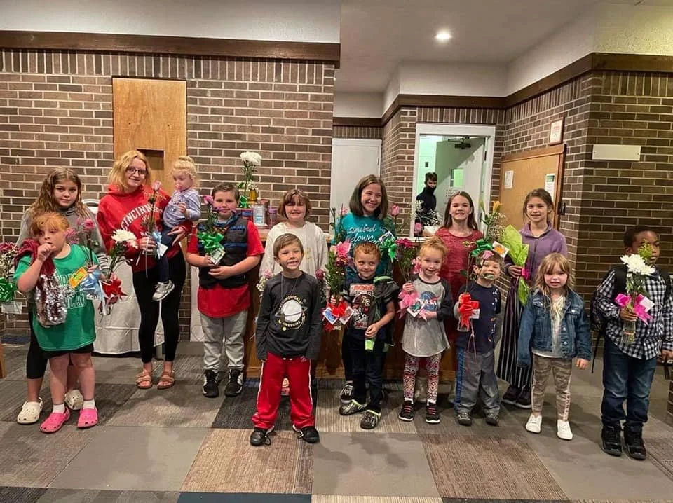 Group of children and an adult holding flowers and gifts inside a brick-walled room.