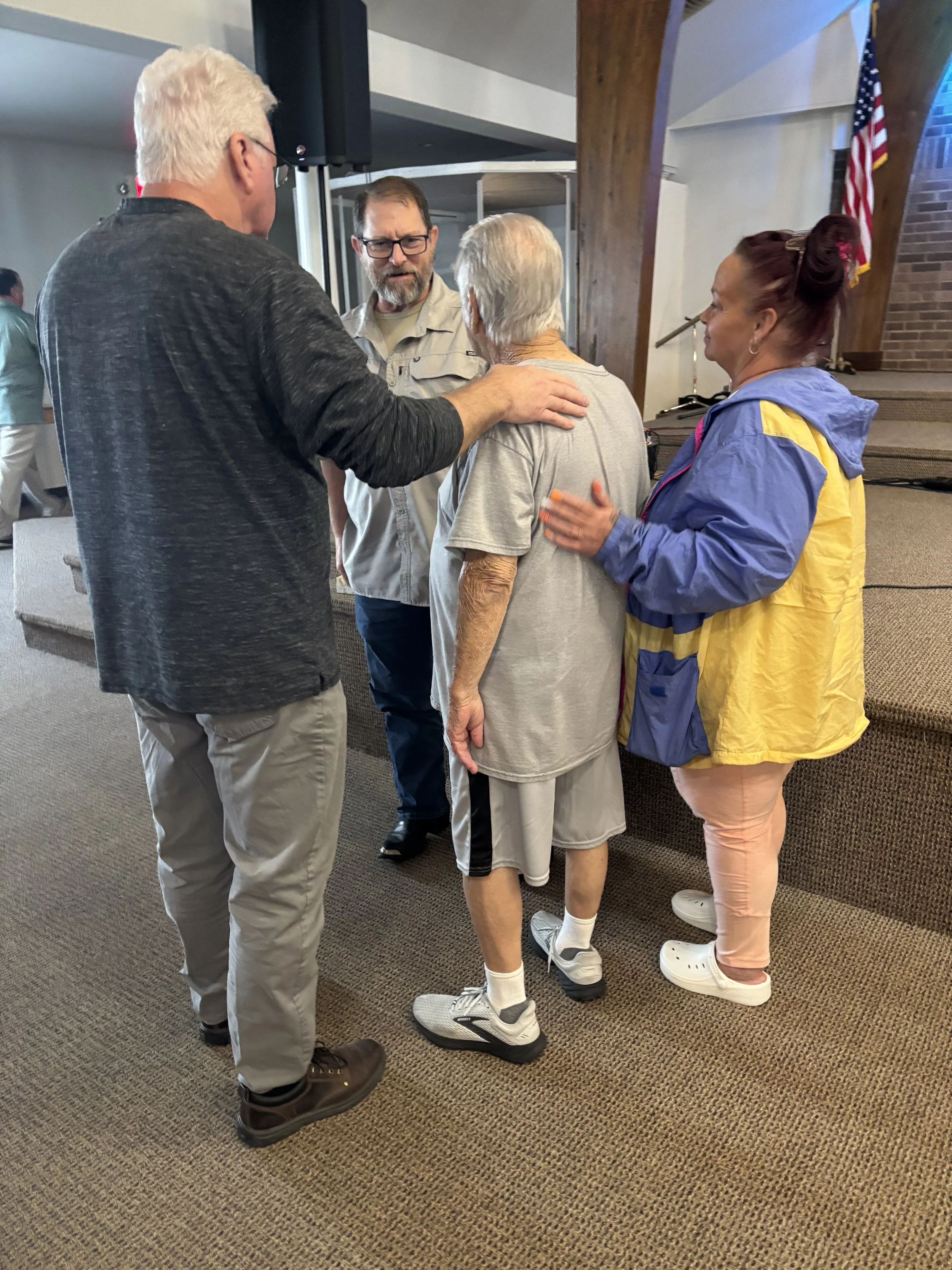 Four people standing in a circle indoors, engaging in conversation. An elderly woman with white hair and athletic clothing is at the center, with three adults surrounding her, one man with gray hair and glasses, another man with dark hair, and a woman with red hair tied up, wearing a yellow and blue jacket. The background features casual, indoor decor including an American flag.