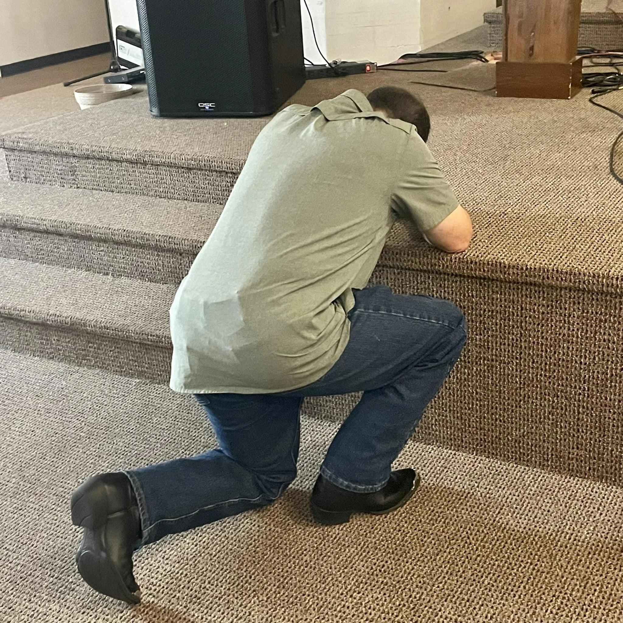 A person kneeling on a carpeted floor with their head bowed, facing away from the camera, wearing a light green shirt and dark blue jeans.