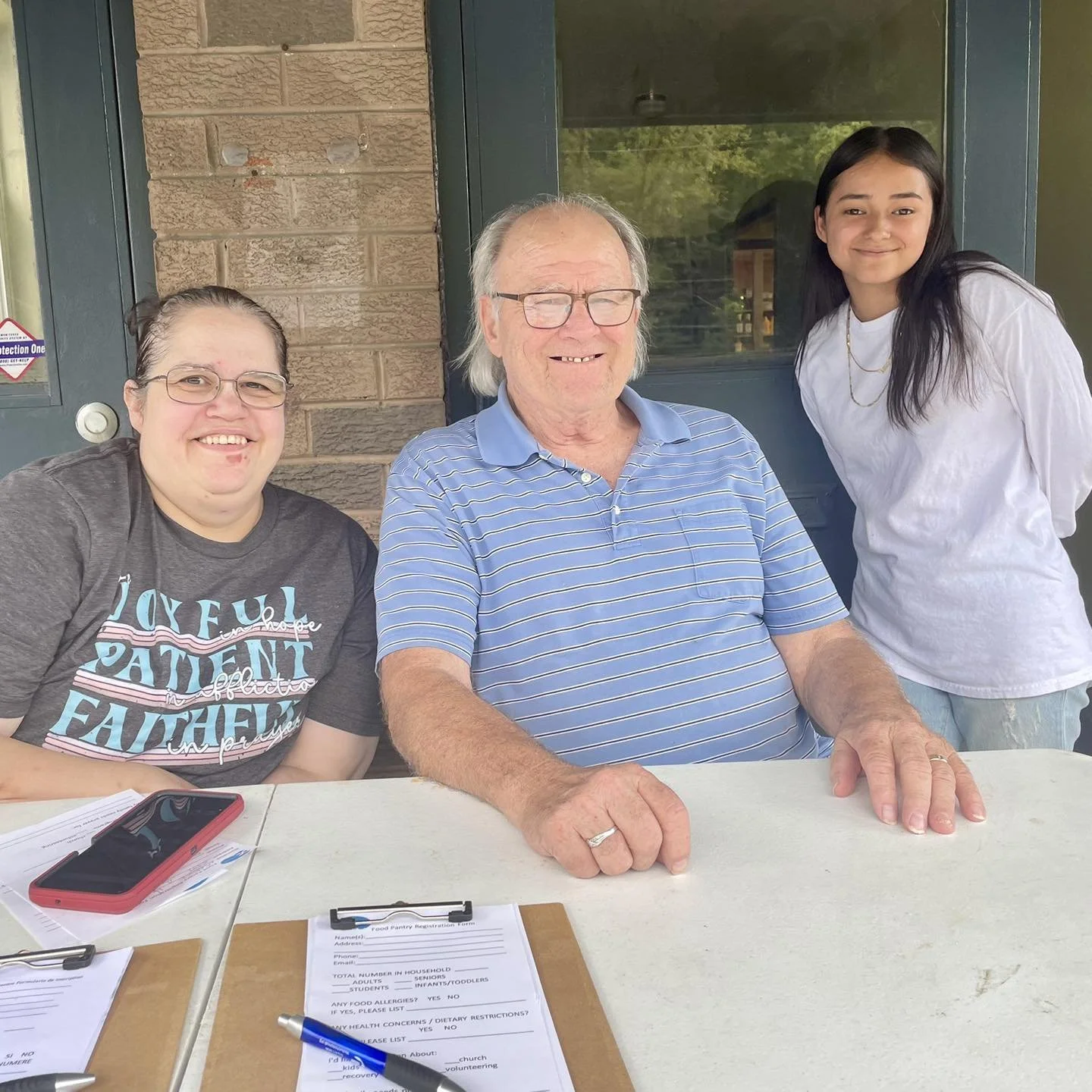 Four people sitting at a table outdoors, smiling for the photo. One man is in the center, with gray hair and glasses, wearing a striped blue shirt. To his left, a woman with glasses and a gray T-shirt. To his right, a young woman with long dark hair and a white shirt. The table has papers, a pen, and a smartphone on it.