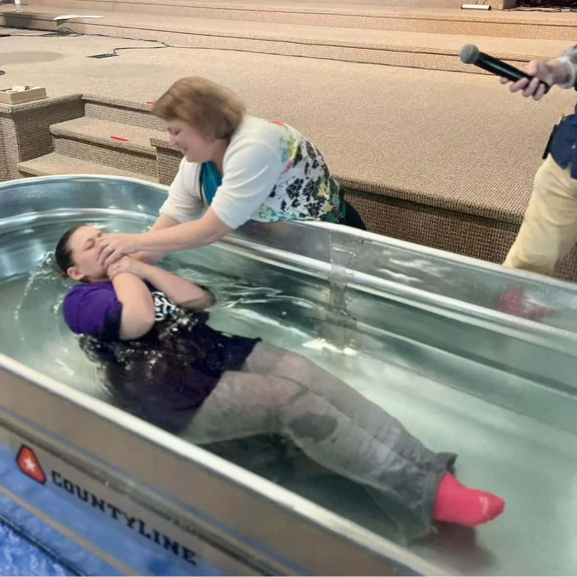 A woman is helping a young girl who is in a small water tub, likely during a religious baptism. The woman is holding the girl's face and shoulders, and the girl appears to be emotional or crying. The scene is indoors on a carpeted floor with a microphone in the background.