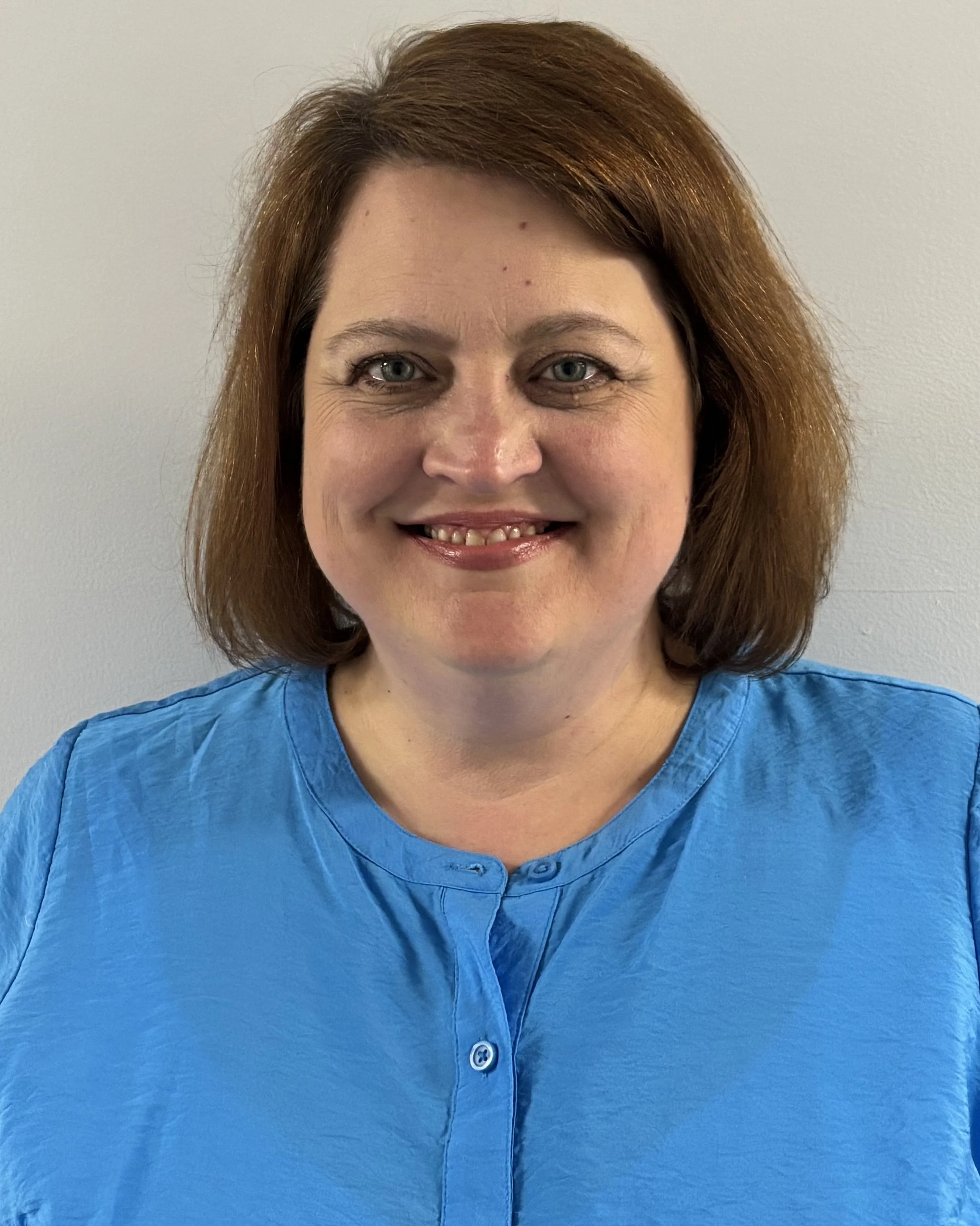 Close-up of a smiling woman with brown hair and blue eyes wearing a blue button-up shirt, standing against a plain beige wall.