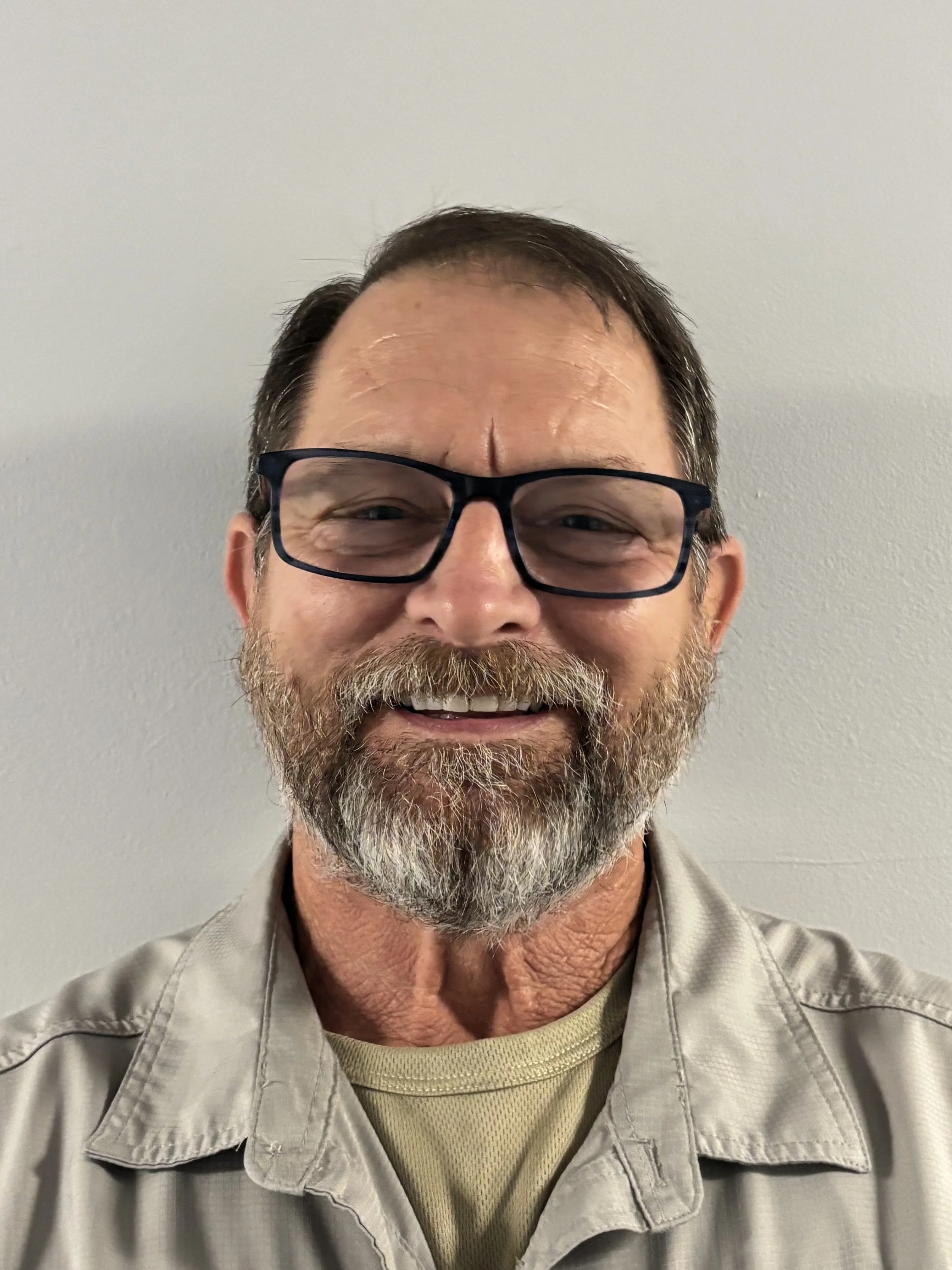 A smiling middle-aged man with glasses, a beard, and short dark hair, wearing a beige jacket over a yellow shirt, standing against a plain light gray wall.