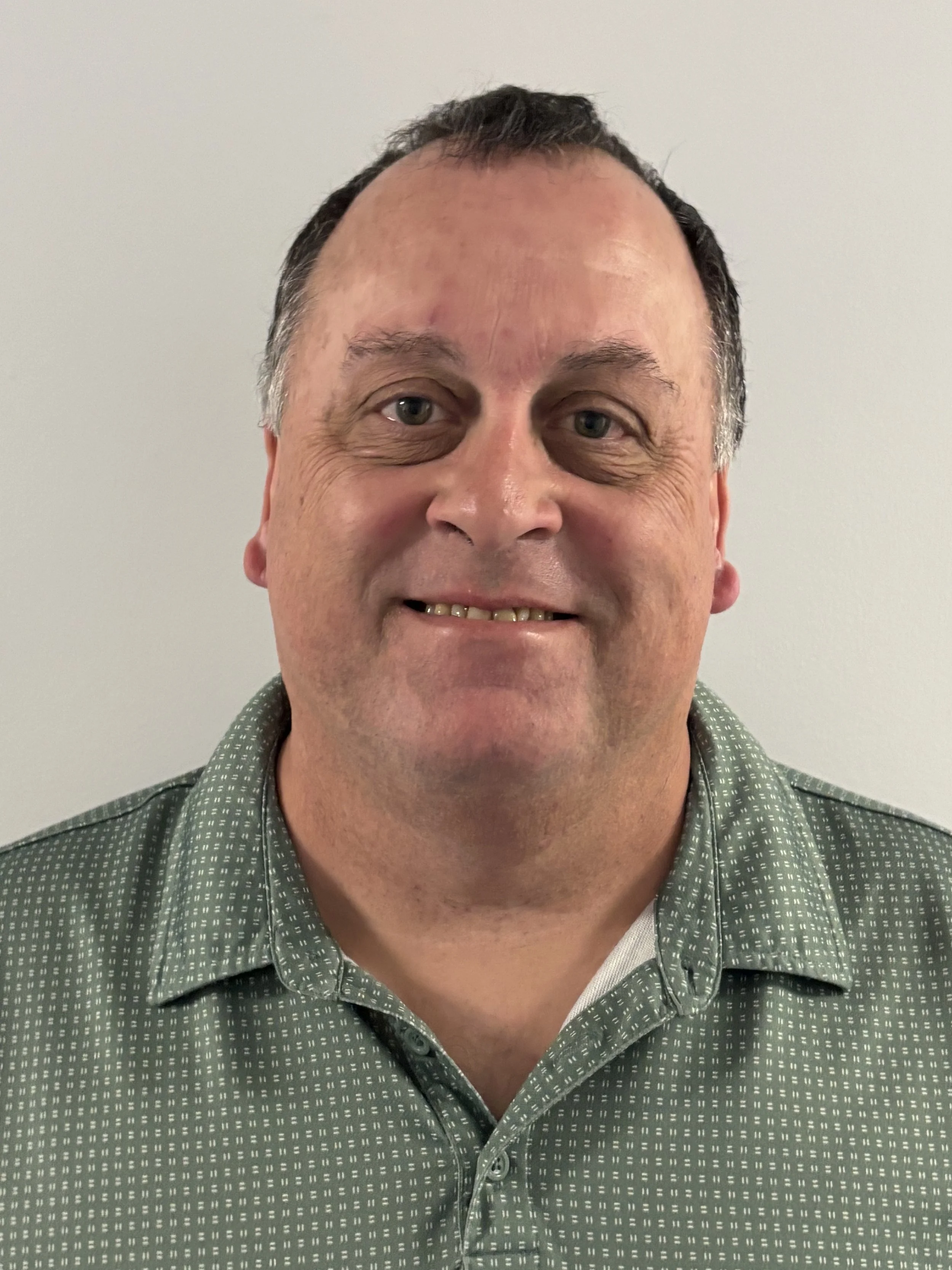 A middle-aged man with short, dark hair, smiling, wearing a green patterned button-up shirt, standing against a plain light gray background.