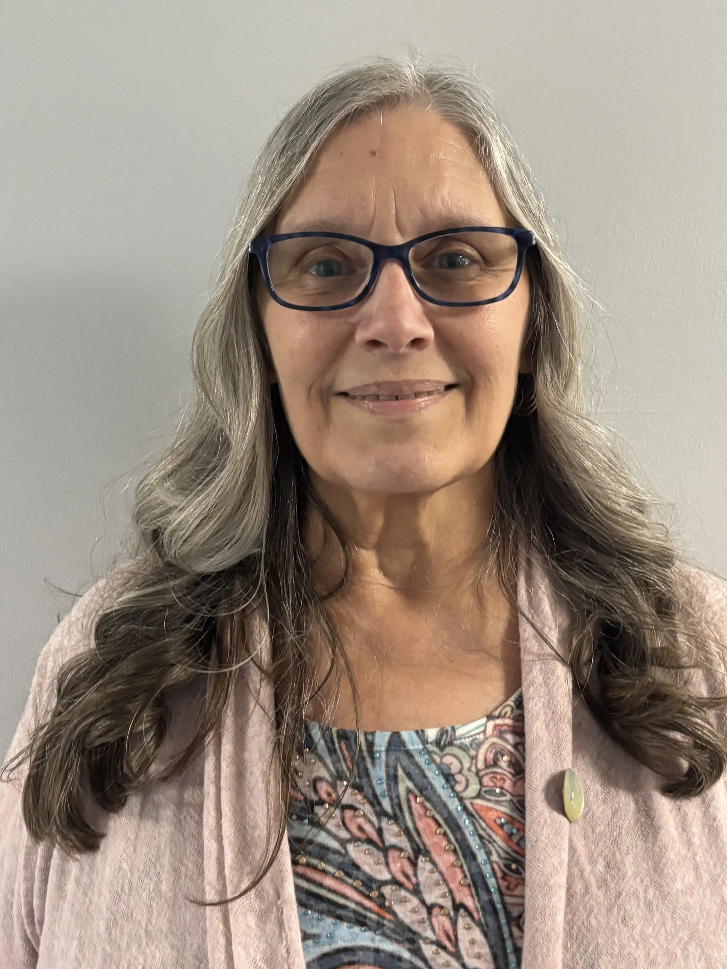 A woman with long, wavy gray hair, glasses, and a light beige coat smiling in front of a plain gray wall.