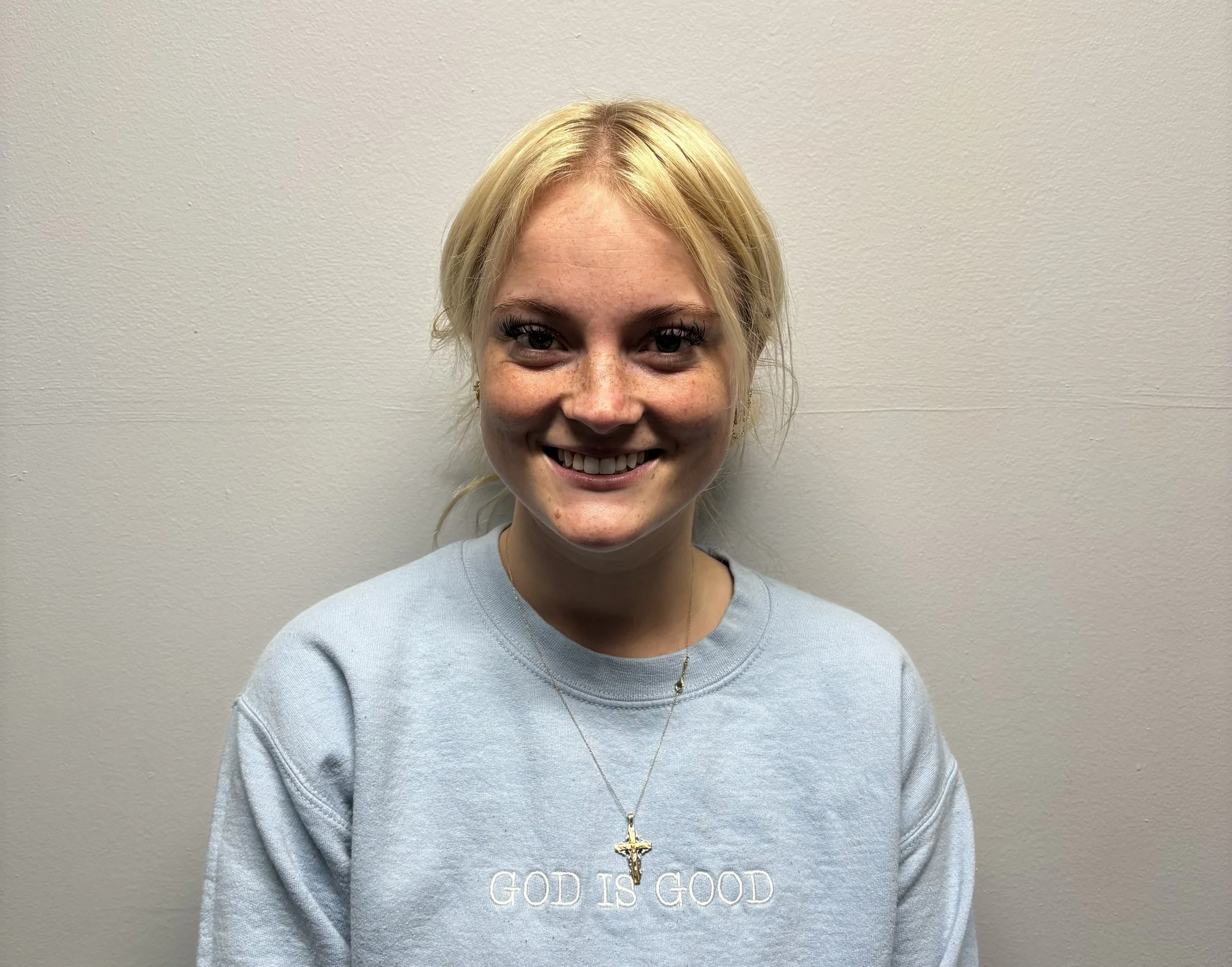 A young woman with blonde hair smiling in front of a plain light-colored wall, wearing a light blue sweatshirt with the text 'GOD IS GOOD' and a gold cross necklace.