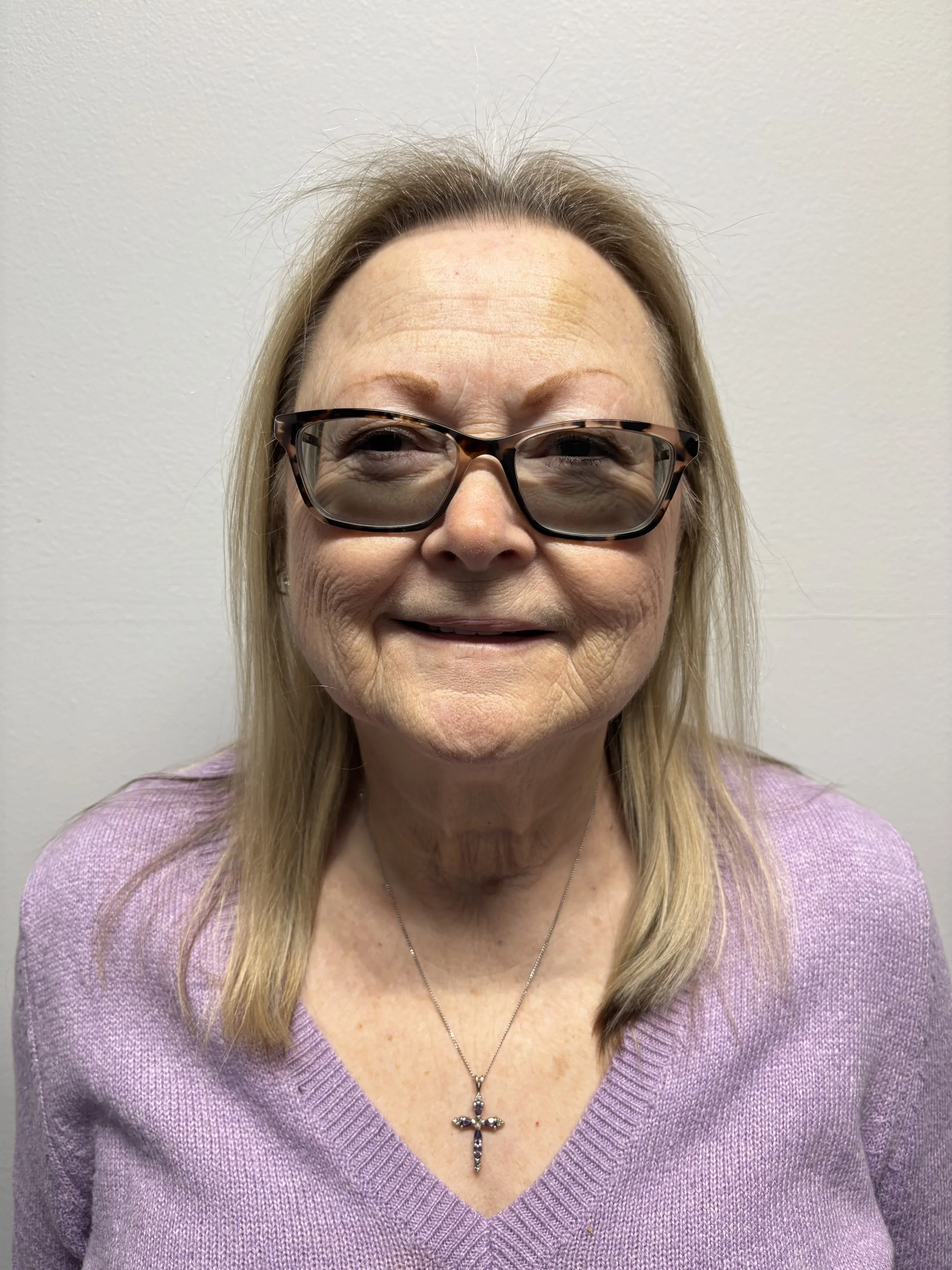A middle-aged woman with blonde hair and glasses, wearing a purple sweater and a cross necklace, smiling against a plain white wall.