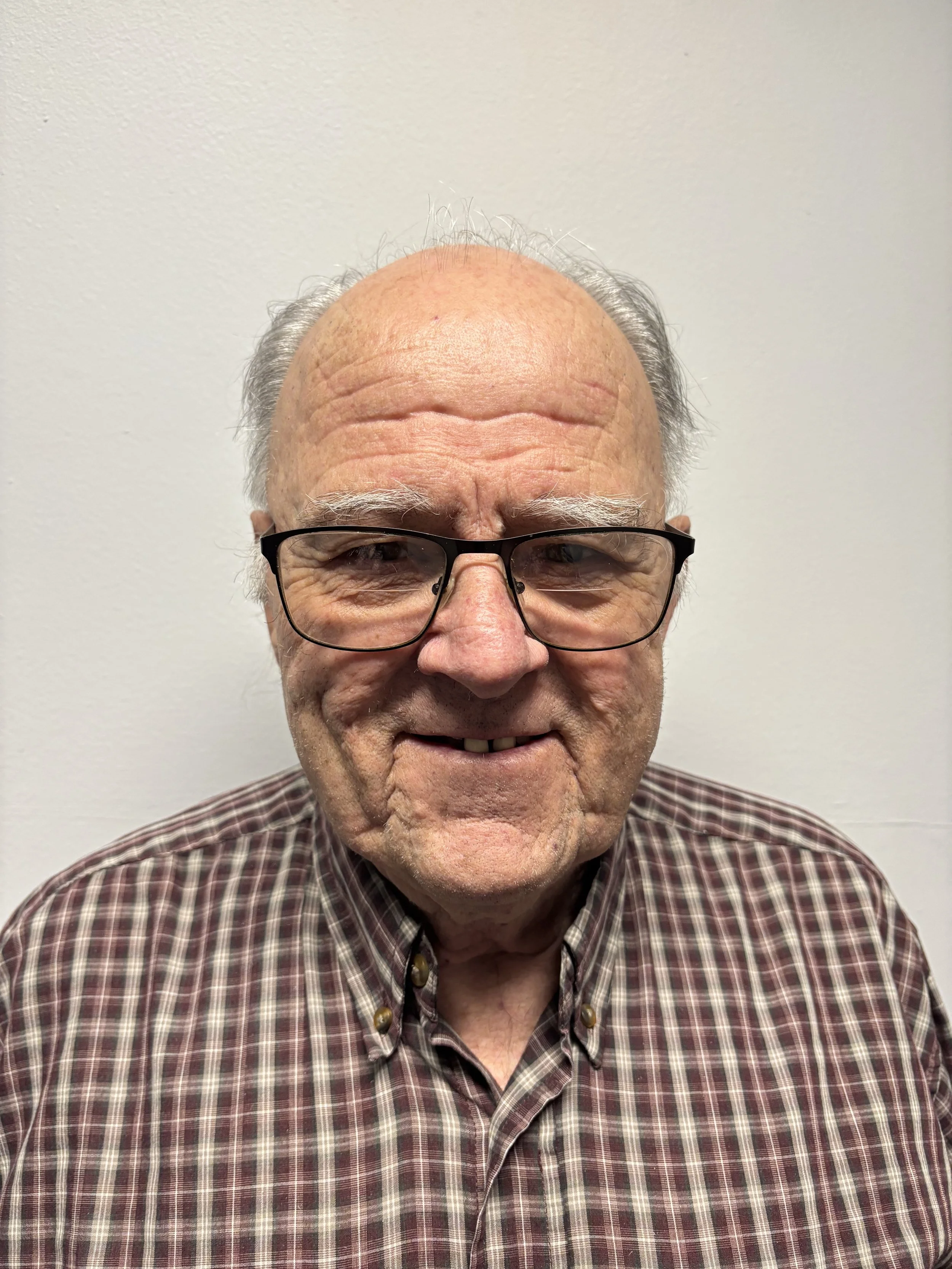 A close-up of an elderly man with gray hair , wearing glasses and a checkered shirt, smiling slightly, against a plain white wall background.