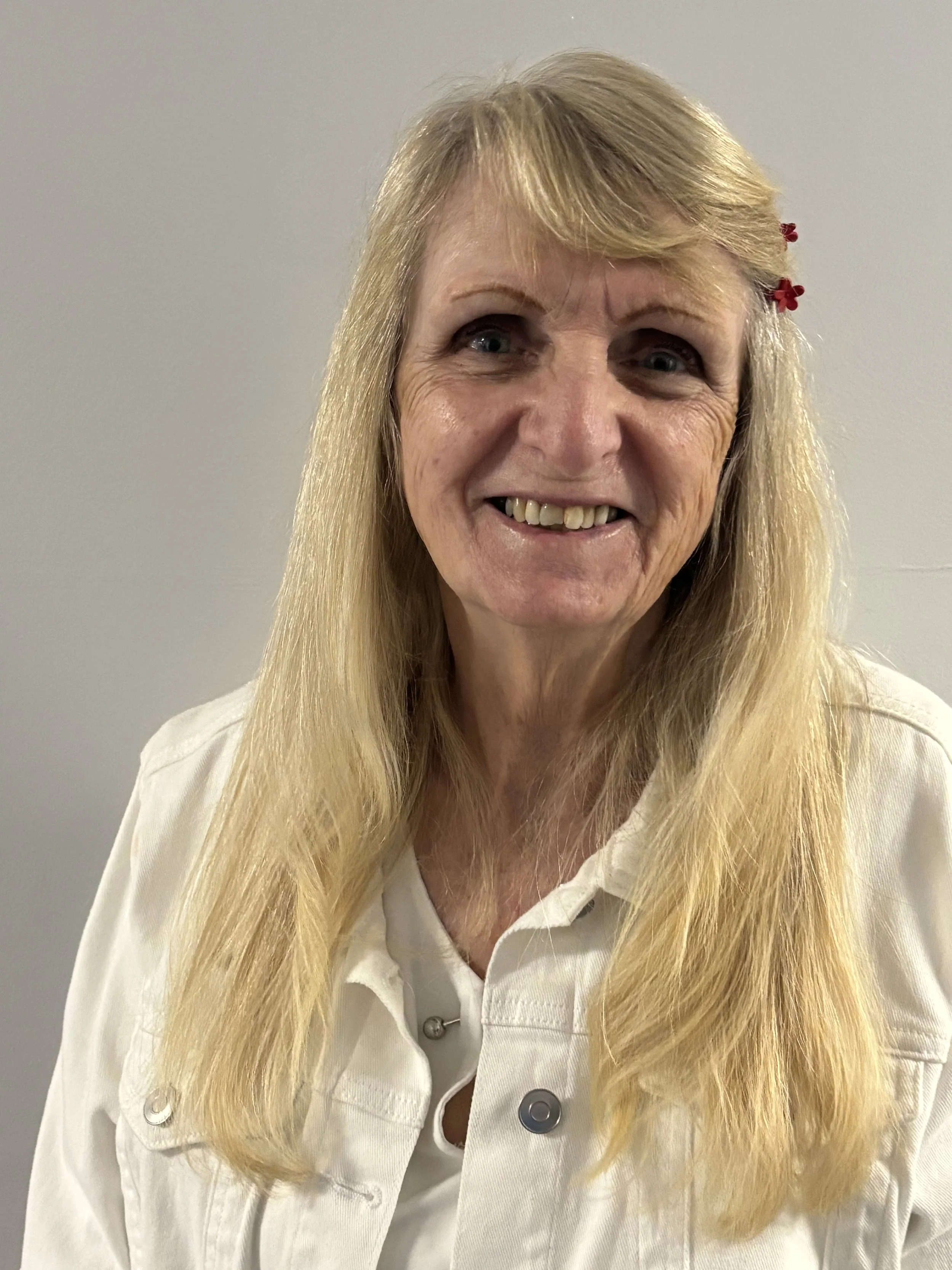 A woman with long blonde hair, blue eyes, and a warm smile wearing a white button-up shirt standing against a plain light gray background.
