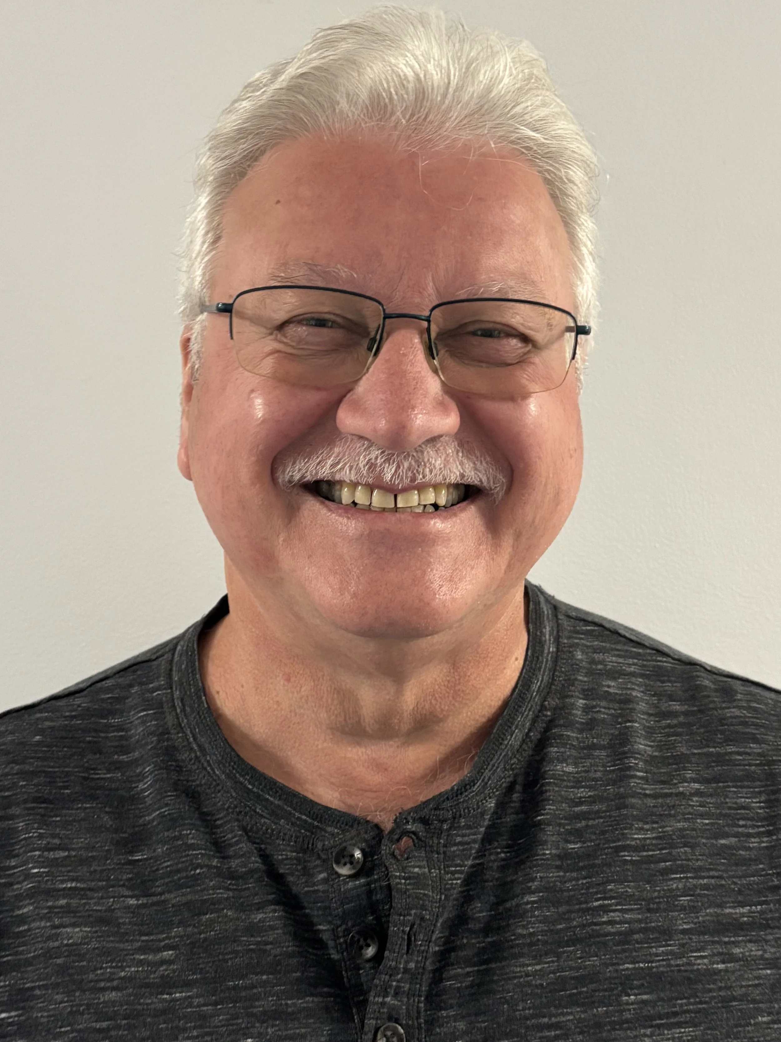 Close-up of smiling middle-aged man with gray hair, glasses, and a dark gray shirt against a plain light background.