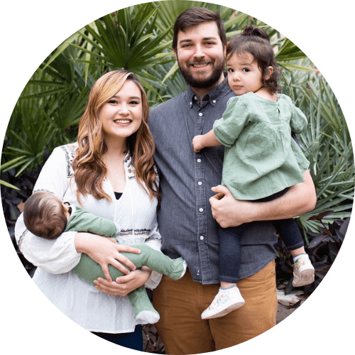 A smiling family of four standing outdoors among green plants, with a man holding a young girl and a woman holding a baby.