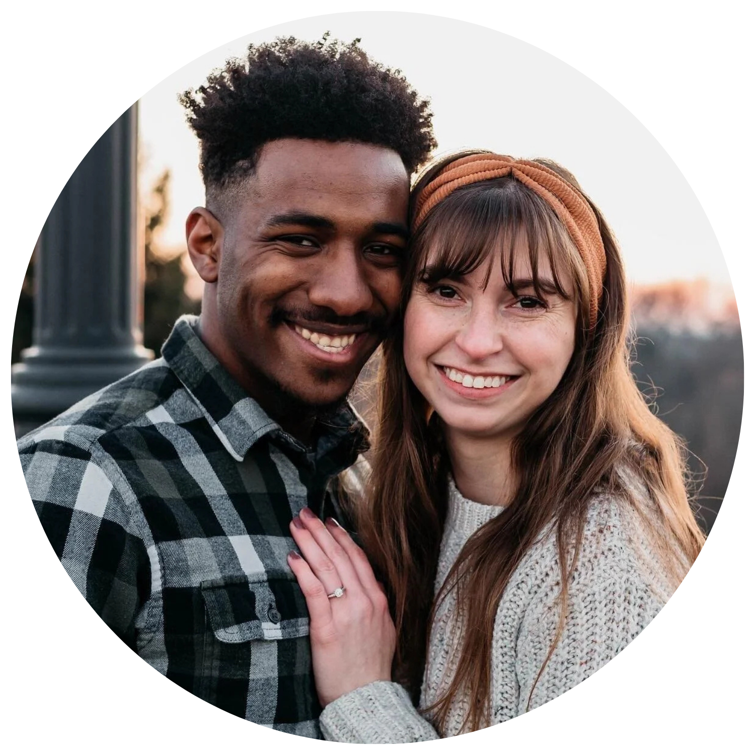 A young couple smiling and embracing outdoors during sunset, with the woman wearing a headband and a knitted sweater, and the man wearing a checkered shirt.
