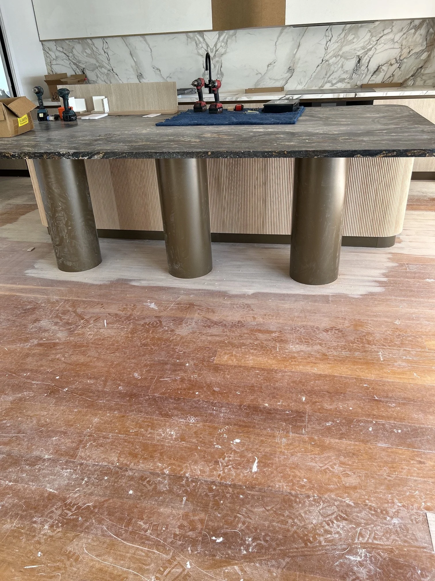 A kitchen island with a black marble countertop and cylindrical metal legs, with tools and materials for construction or renovation. In the background, a marble backsplash and wooden and white cabinetry are visible.