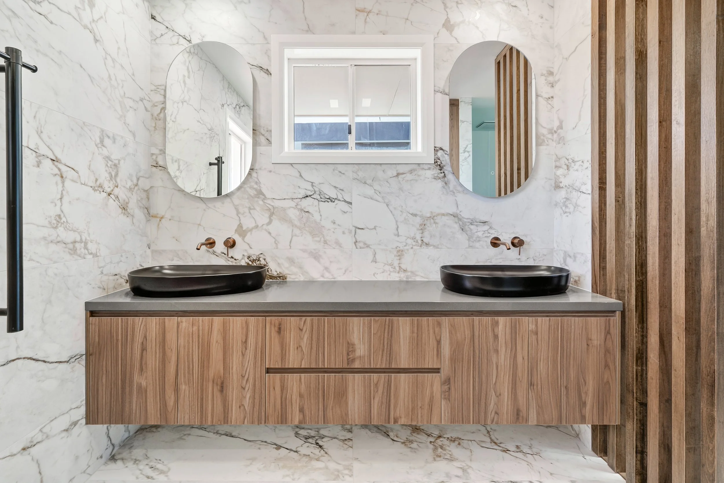 Modern bathroom with marble walls and floor, featuring a double vanity with wood cabinetry and black vessel sinks, large oval mirrors, a window above the vanity, and a wooden slatted wall on the right.