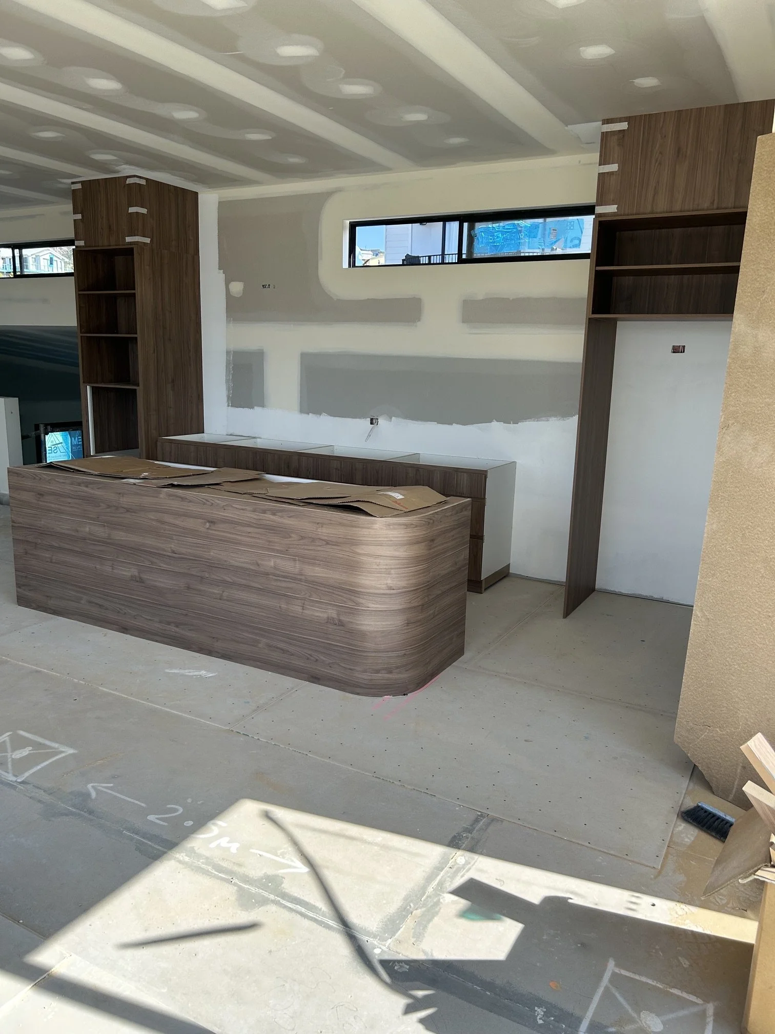 Interior of a building under construction with a curved wooden reception desk, wooden bookshelves, and a partially finished wall.
