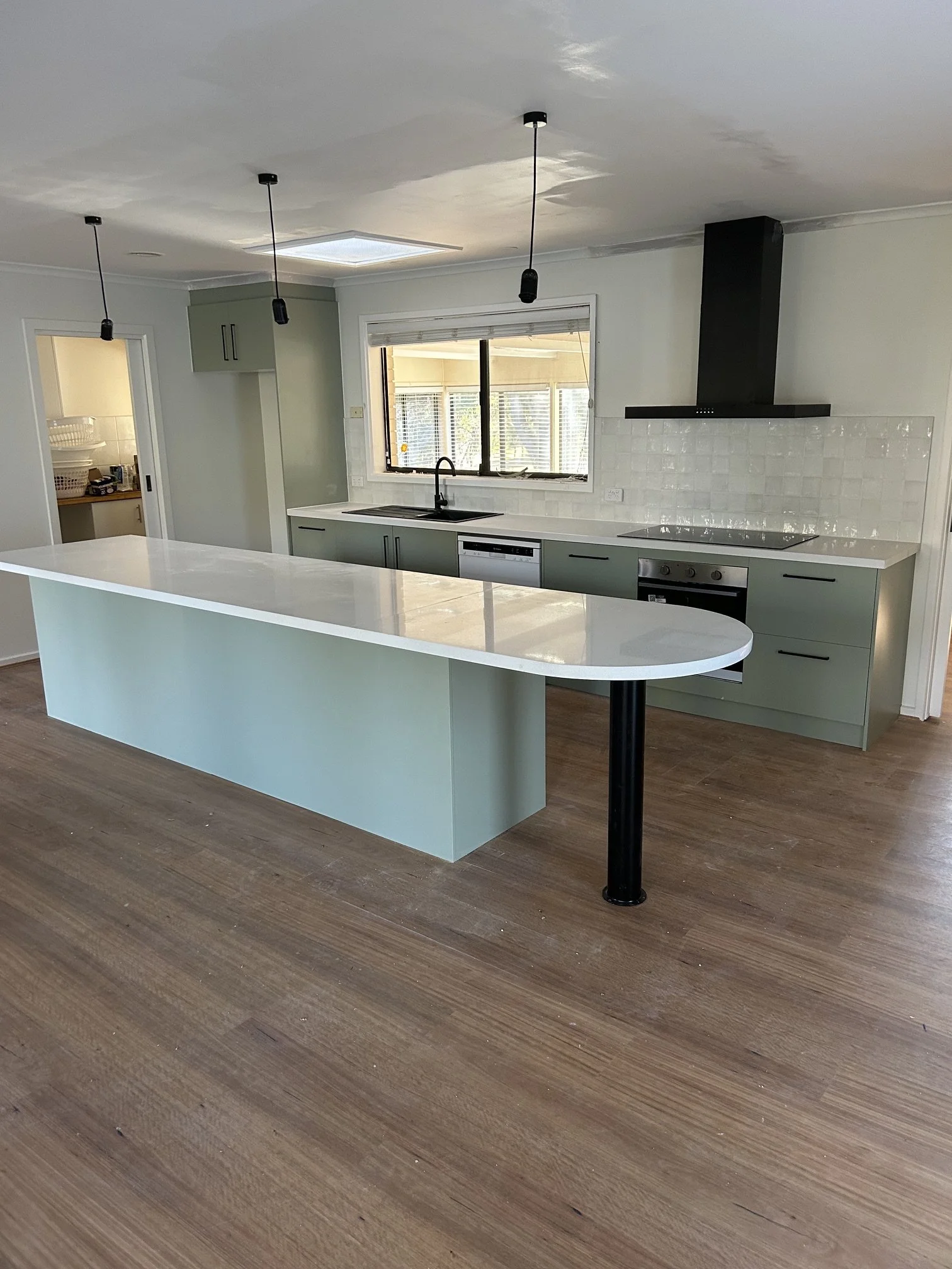 Modern kitchen with mint green cabinets, white countertops, and a black range hood. A large window above the sink lets in natural light. There is an island with a mint green base and a white countertop, and two black pendant lights hang from the ceiling. The floor is wood.
