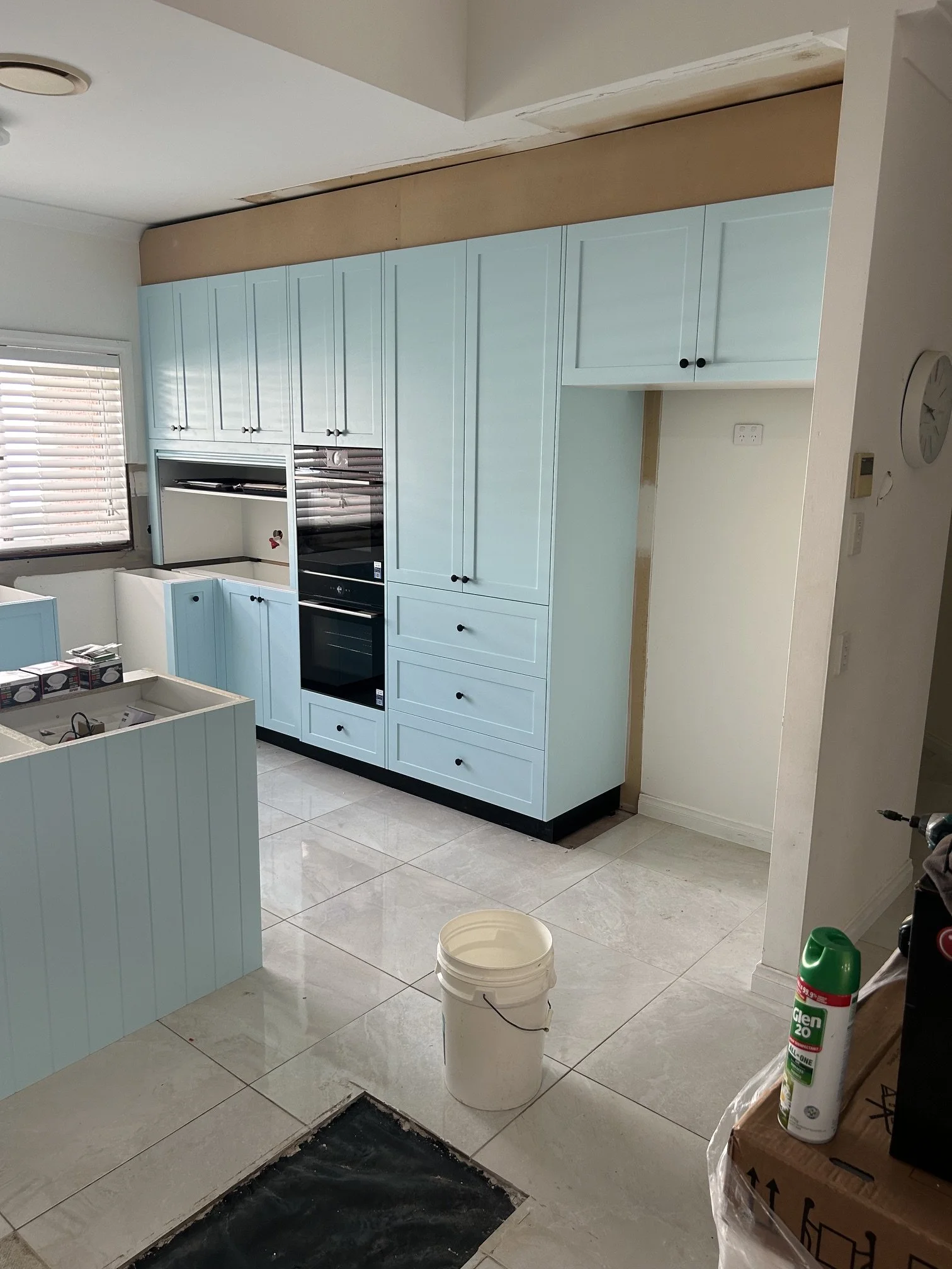 A kitchen under renovation with light blue cabinets and a black oven. The floor is tiled, and there is a window with blinds. Construction tools and materials, including a bucket, a can of Glem glue, and a cardboard box, are scattered around.