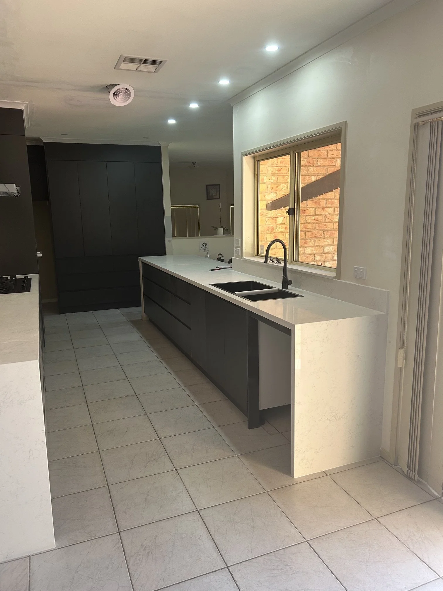 Modern kitchen with white countertops, dark lower cabinets, tile floor, large window showing brick wall outside, and ceiling lights.