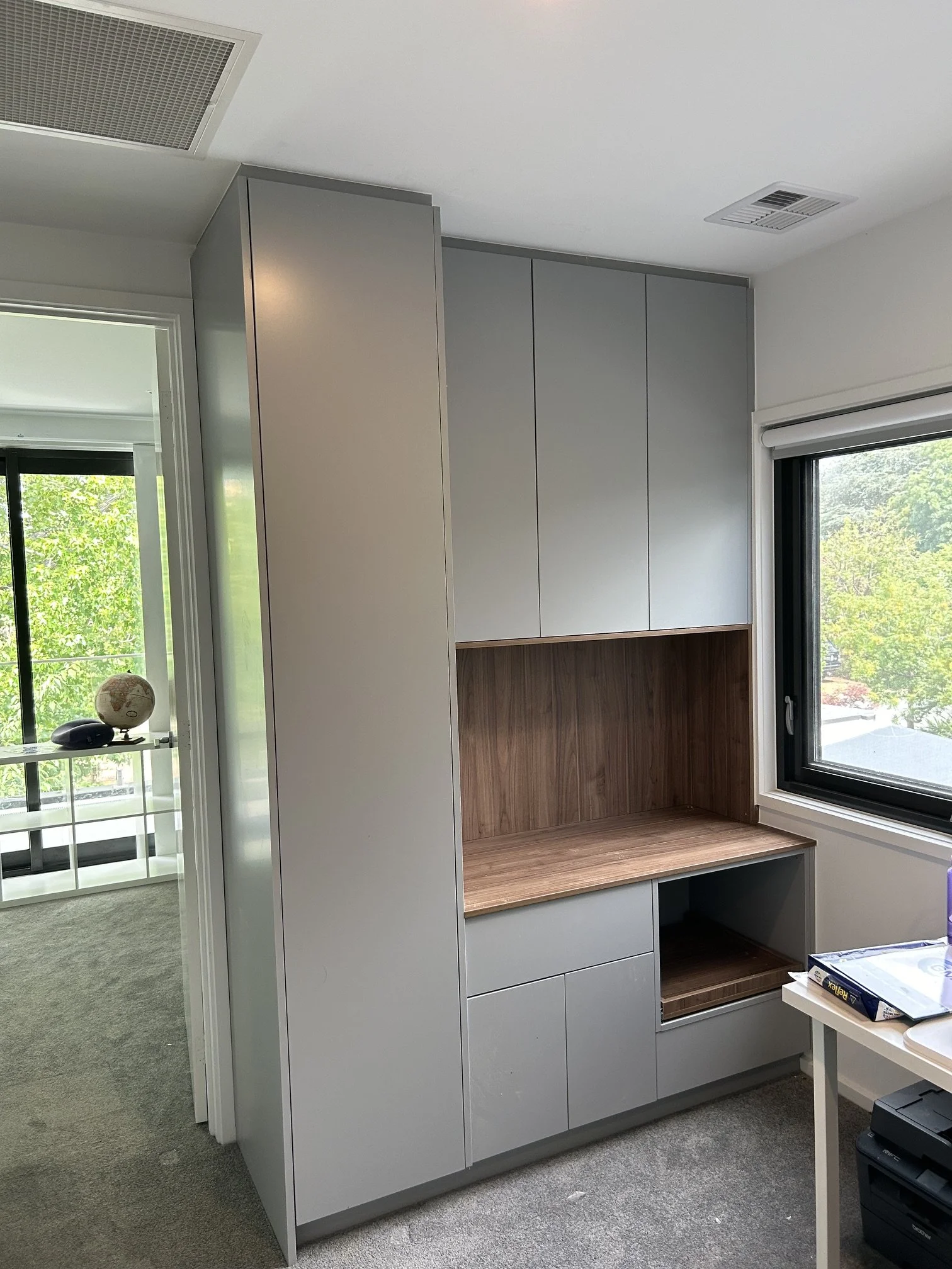 A modern built-in cabinet with closed white doors and an open wooden shelf, positioned next to a window with a view of trees outside, in a room with carpeted floor.