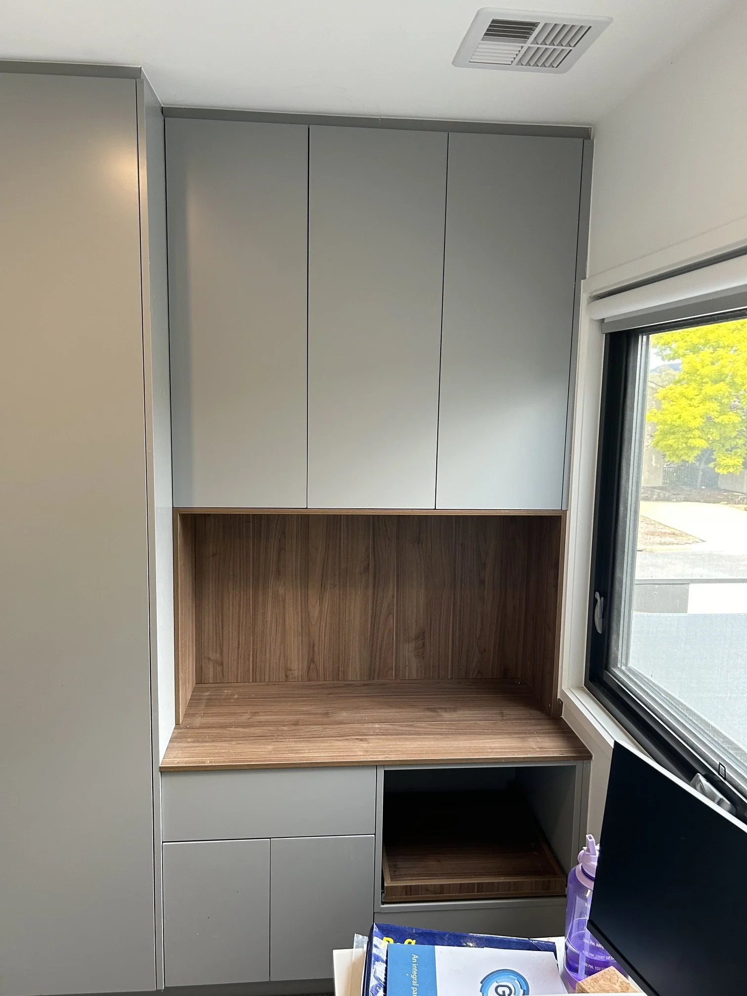 Empty kitchen cabinet with open space at the bottom, above a wooden shelf, next to a window with a view of trees and a street.