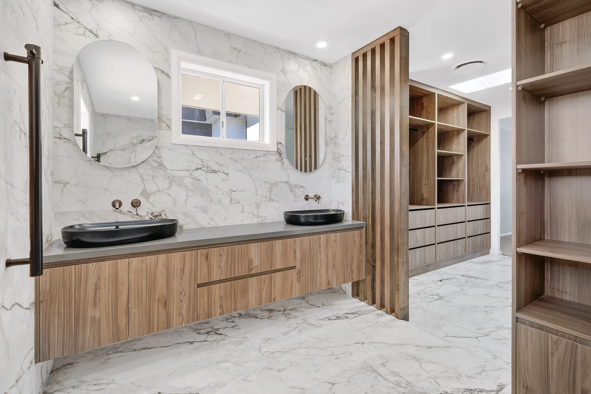 Modern bathroom with marble walls and floor, a double black vessel sink vanity, round mirrors, a window, and wooden shelving units.