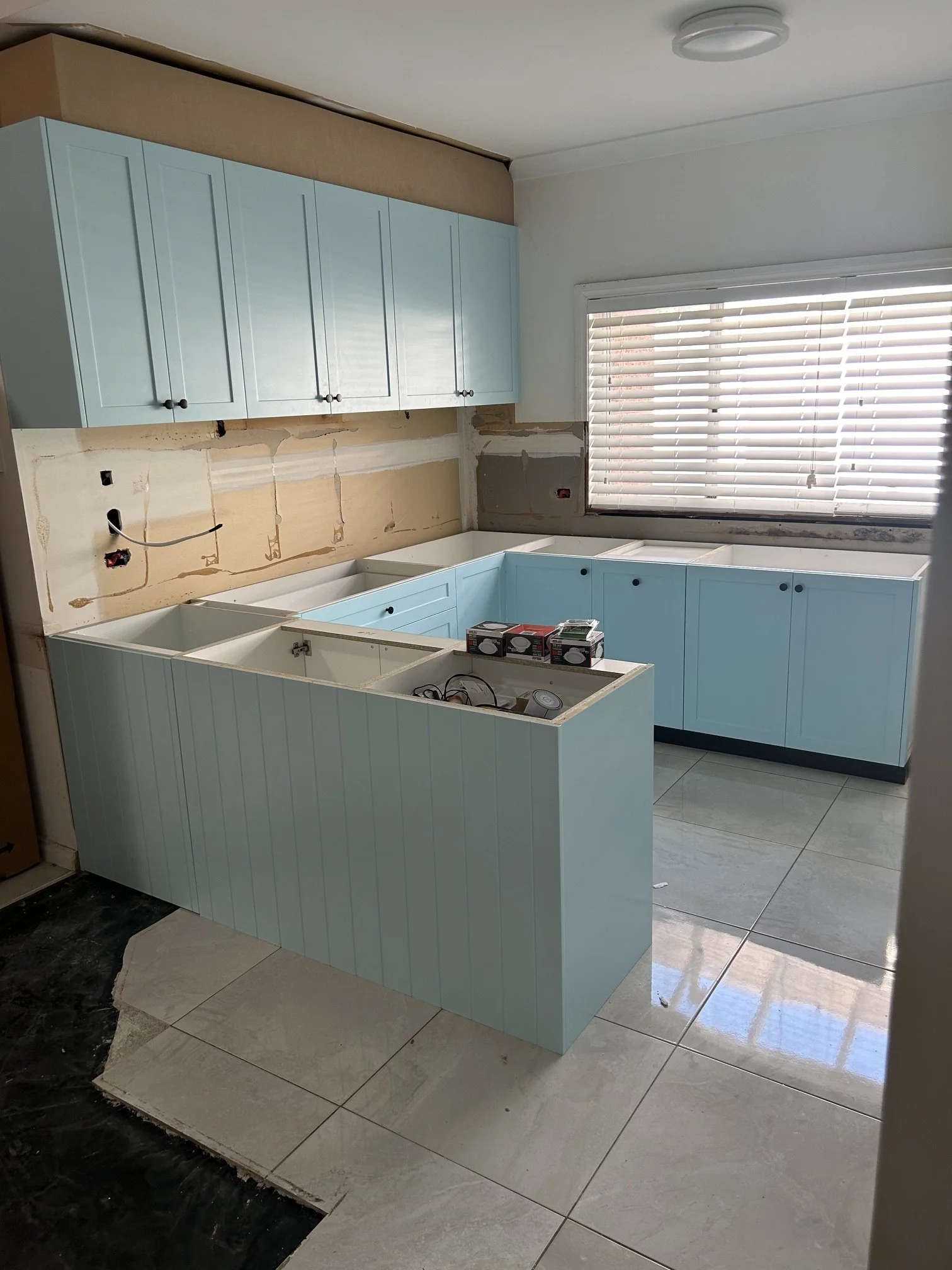 Kitchen under renovation with light blue cabinets installed, some cabinets are missing doors, large window with blinds, and construction tools and materials visible.