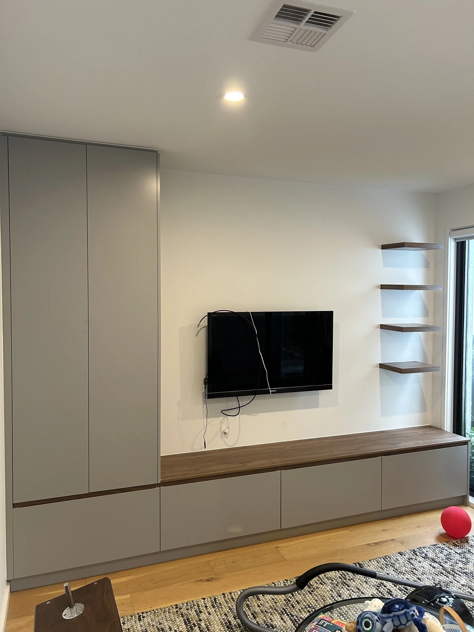 Modern living room with gray cabinetry, wall-mounted black flat-screen TV, floating shelves, a window with blinds, a hardwood floor, and a patterned rug.